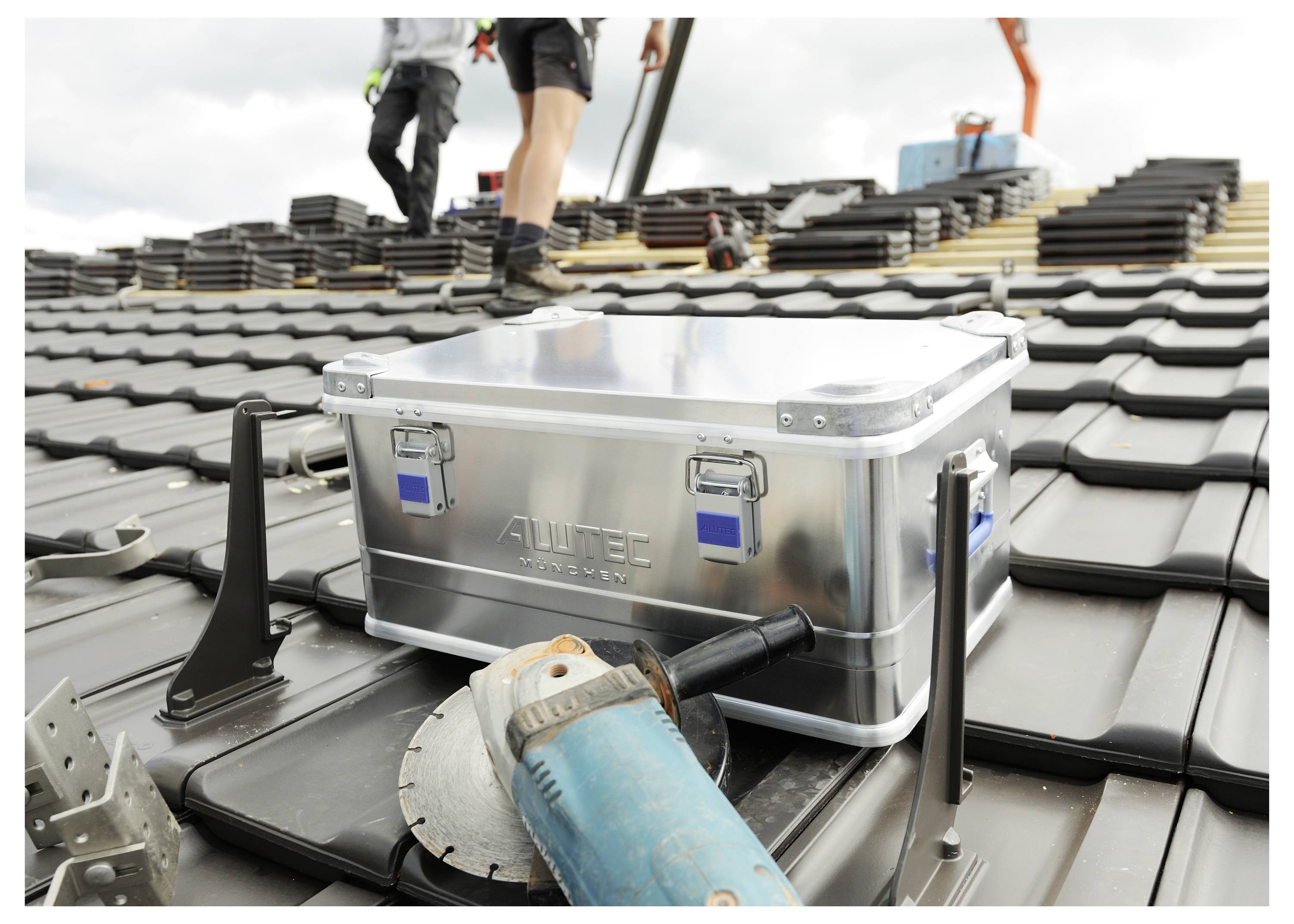 Workers installing tiles on a rooftop with tools and a shiny metal toolbox nearby.