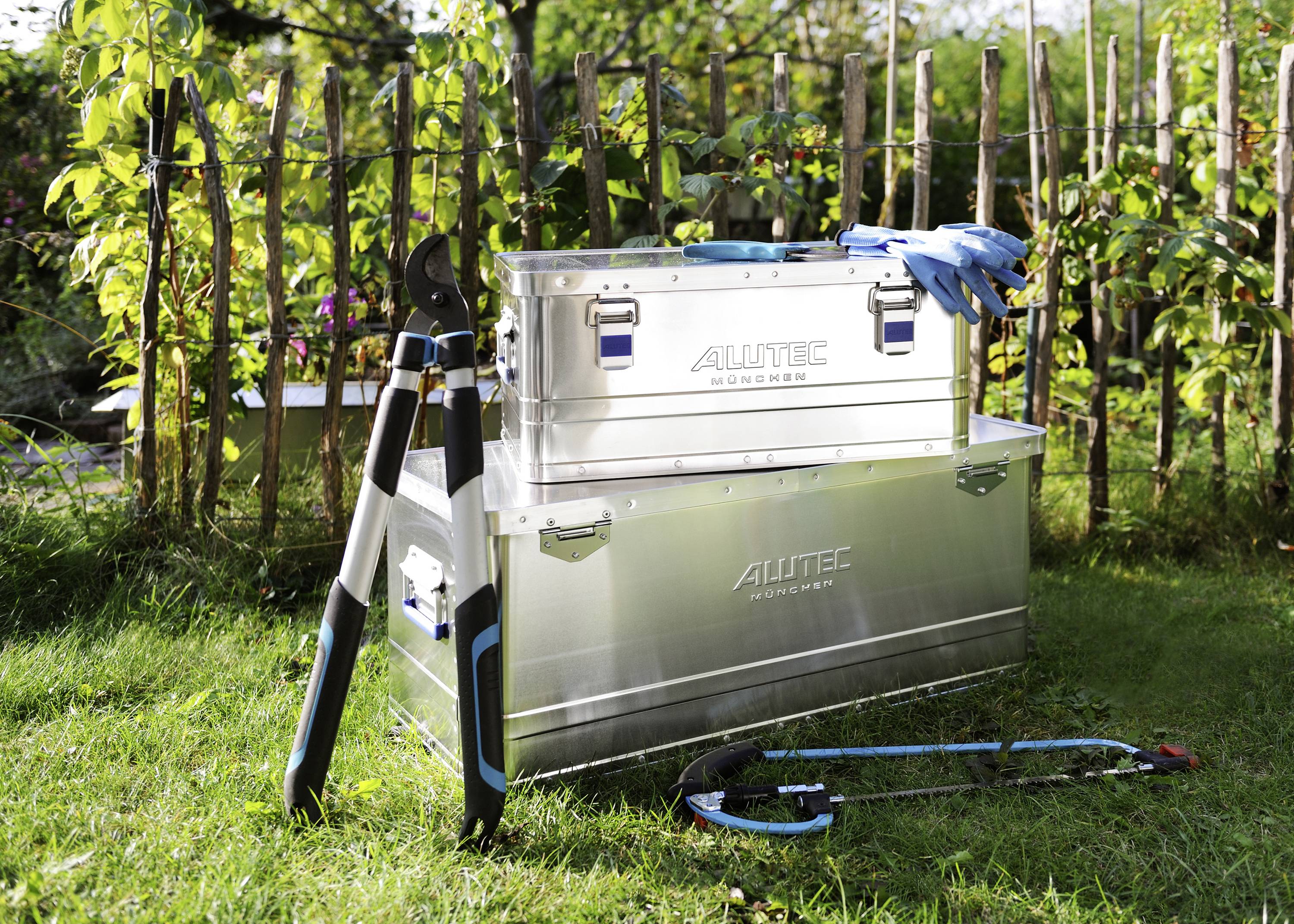 Two silver toolboxes with the inscription 'ALUTEC' are standing in the garden. Next to them, garden shears and a saw blade lie on the grass.