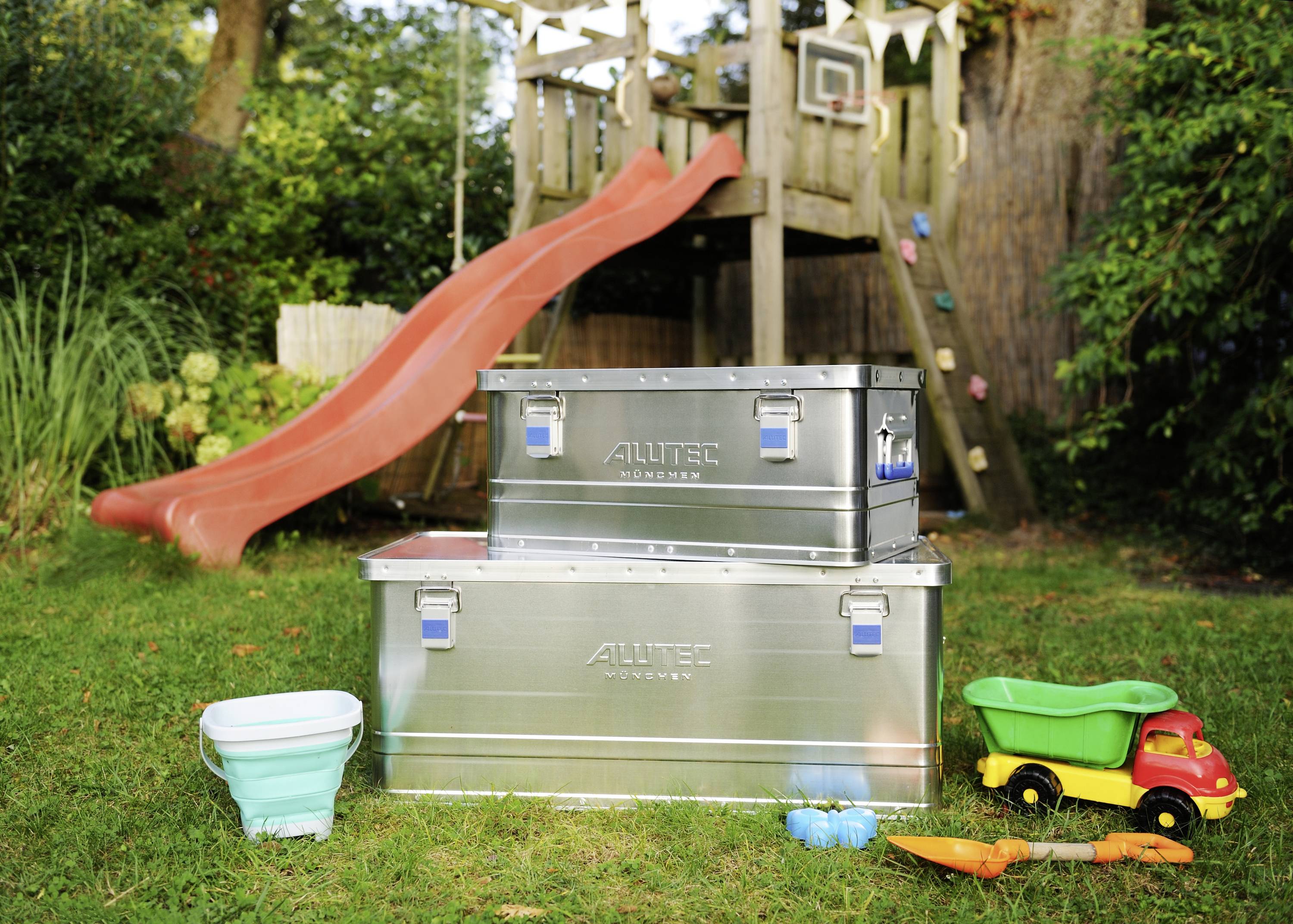 Two metal boxes are standing on a grass verge in front of a playground with a slide. Toys, including a lorry and a bucket, are scattered around.