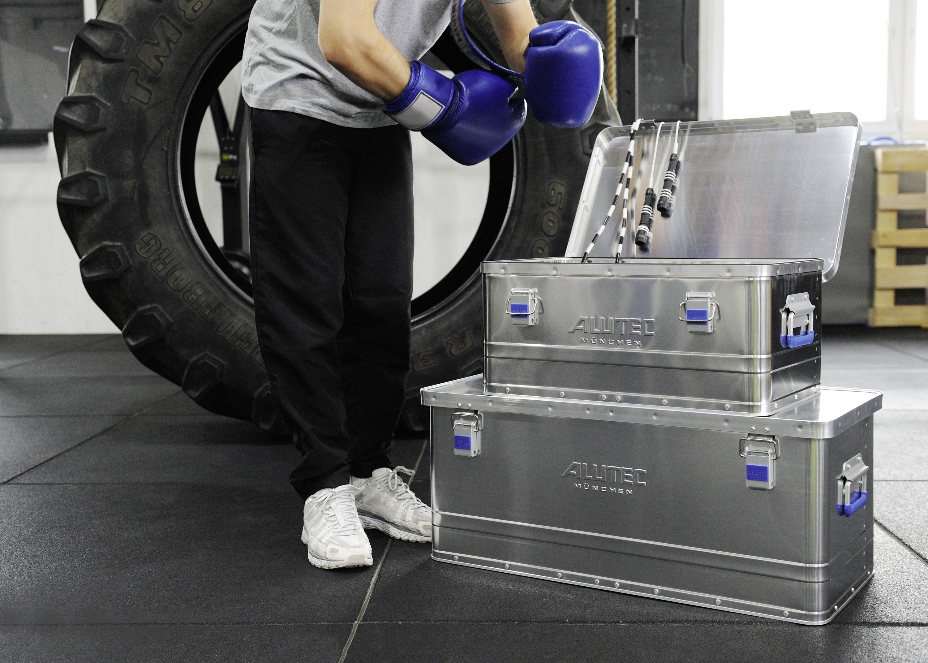 A person wearing sports clothing and boxing gloves stands next to two metal boxes, one of which is open, in a gym in front of a large tyre.