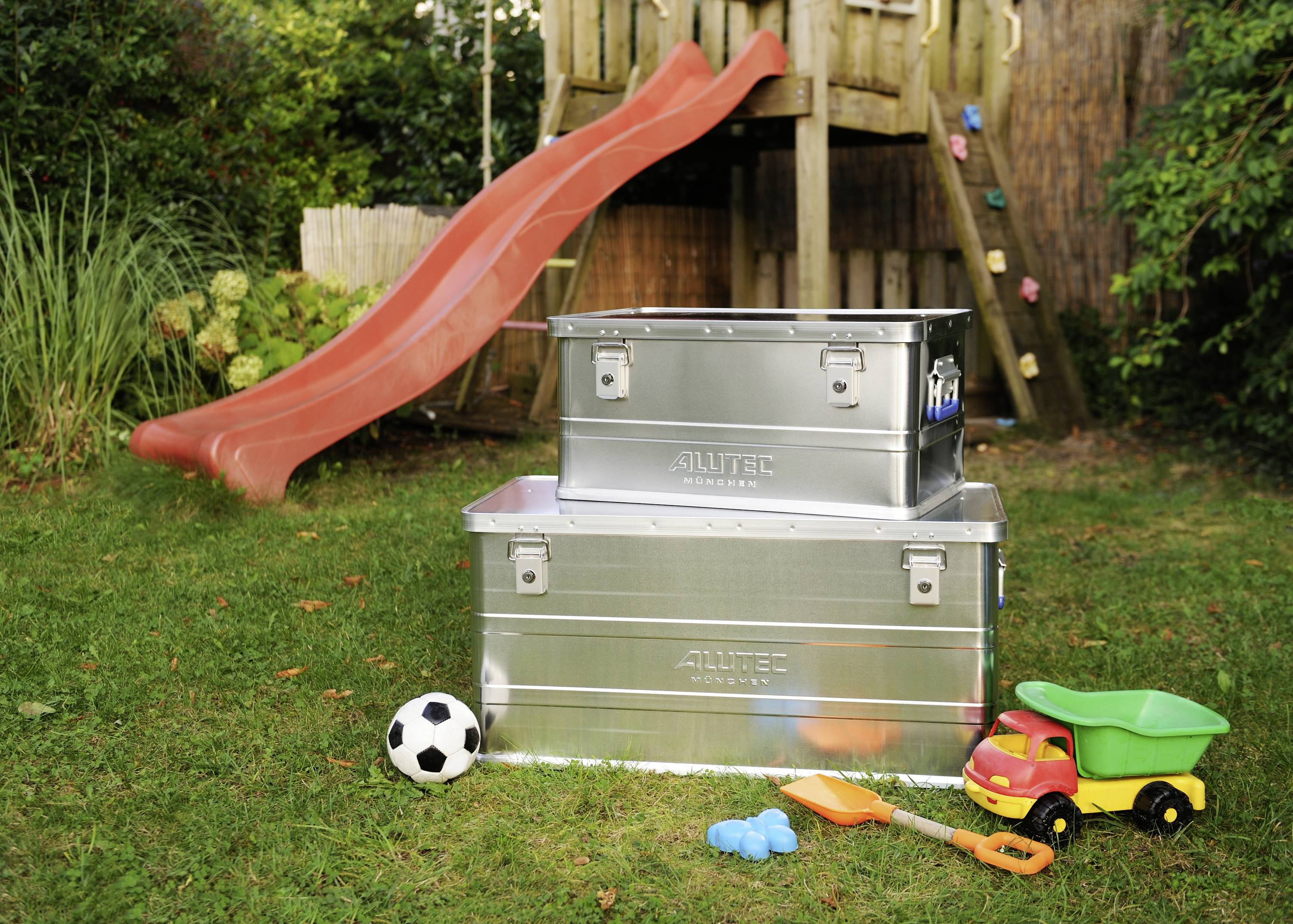 Two metal boxes on a grassy area in front of a red slide, surrounded by children's toys such as a football and a toy car.
