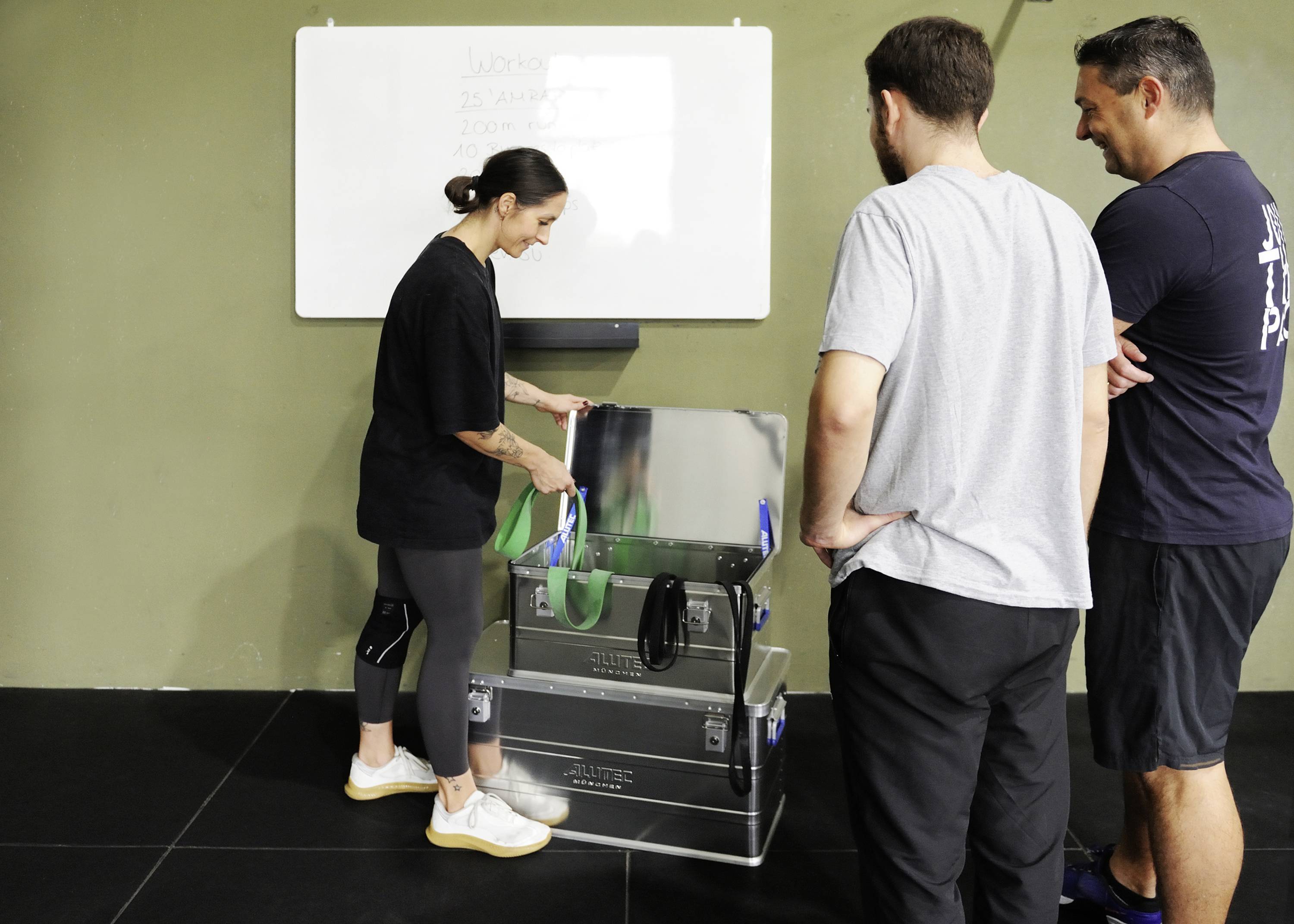 A person is showing something in a box to two other individuals in a gym. A whiteboard is hanging on the wall in the background.