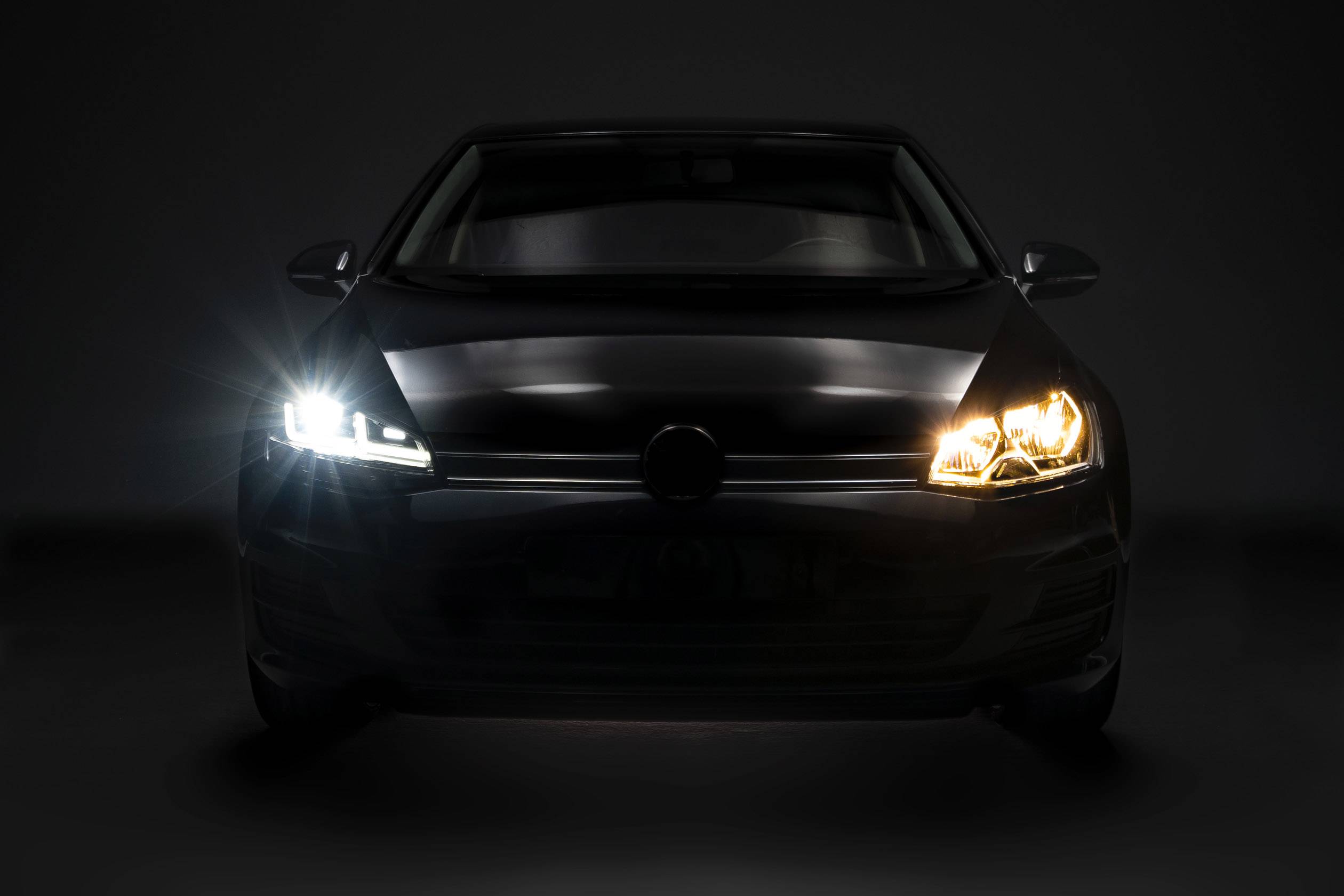 A car is positioned head-on against a dark background. Its headlights are on, with one light shining white and the other orange.