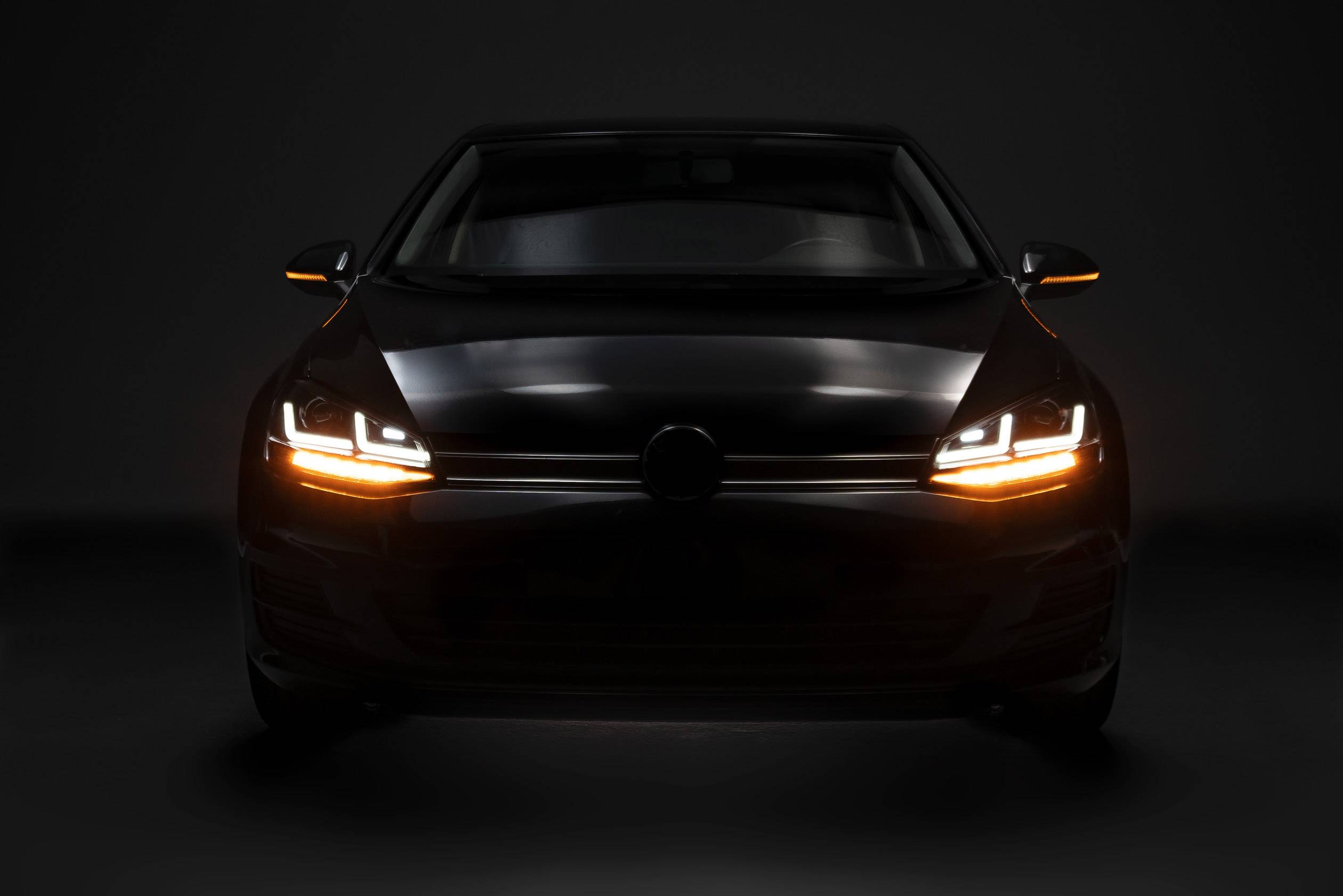 A black car, photographed head-on, with headlights and indicator lights switched on, against a dark background.