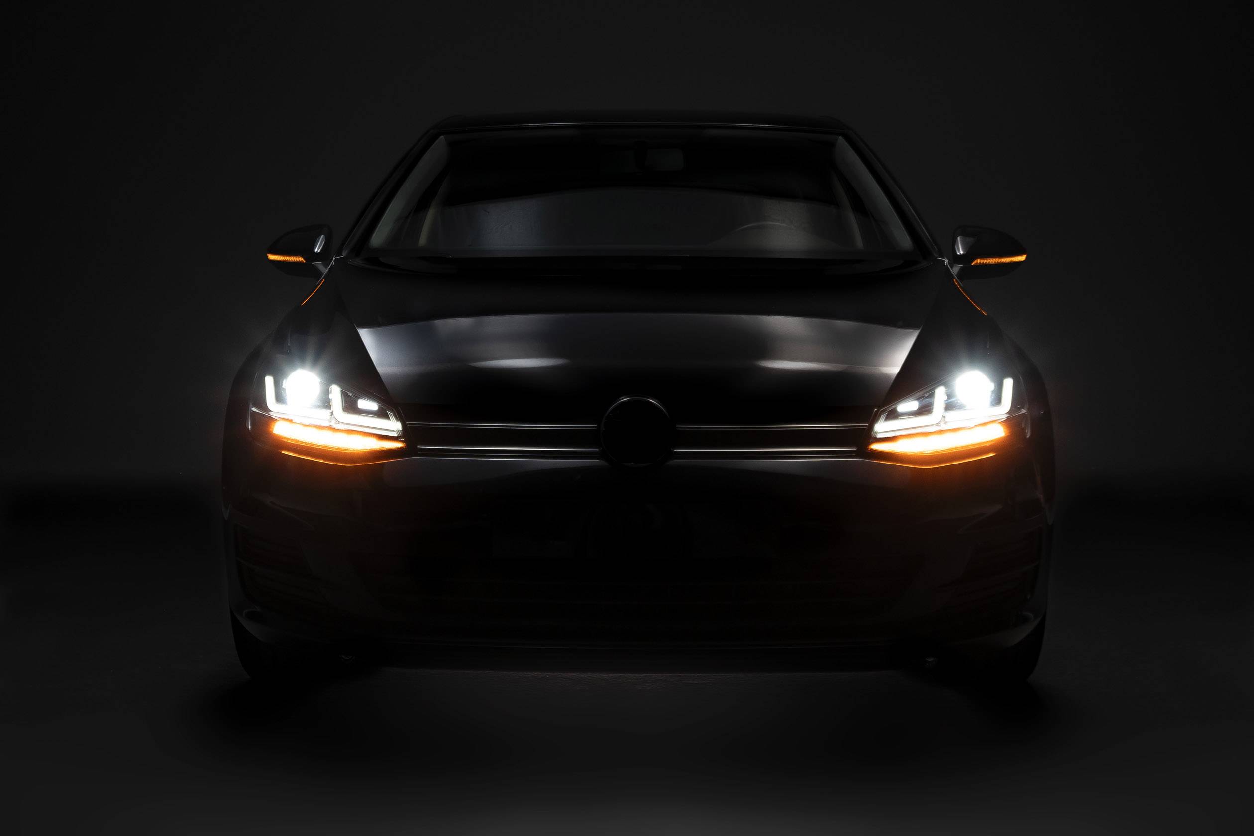 A car from the front, headlights and indicator lights switched on, in a dark environment. The vehicle is the focal point of the image.