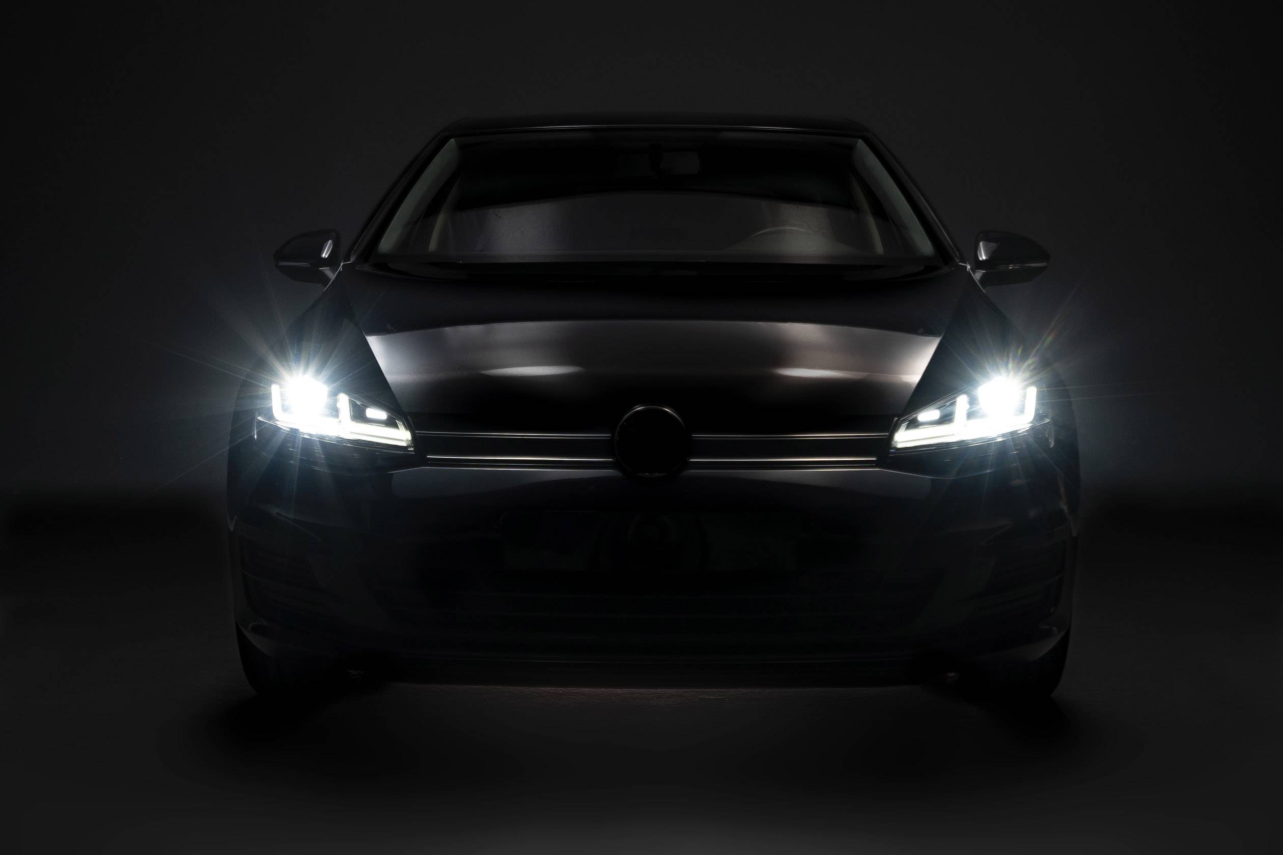 The image shows the front view of a car with dipped headlights against a dark background.
