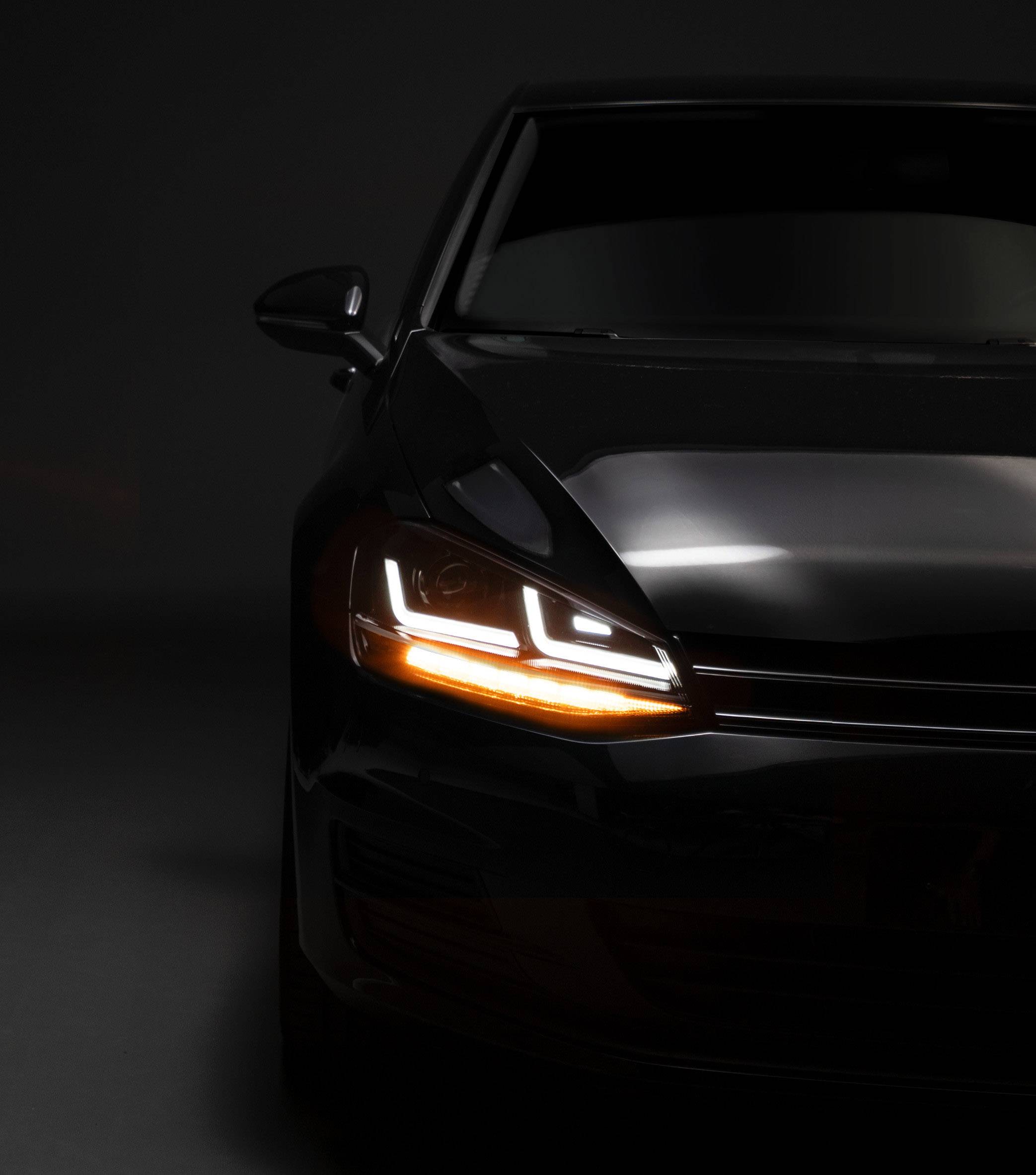 A black car with its headlight switched on stands in a dark room, with only the front section accentuated by light.