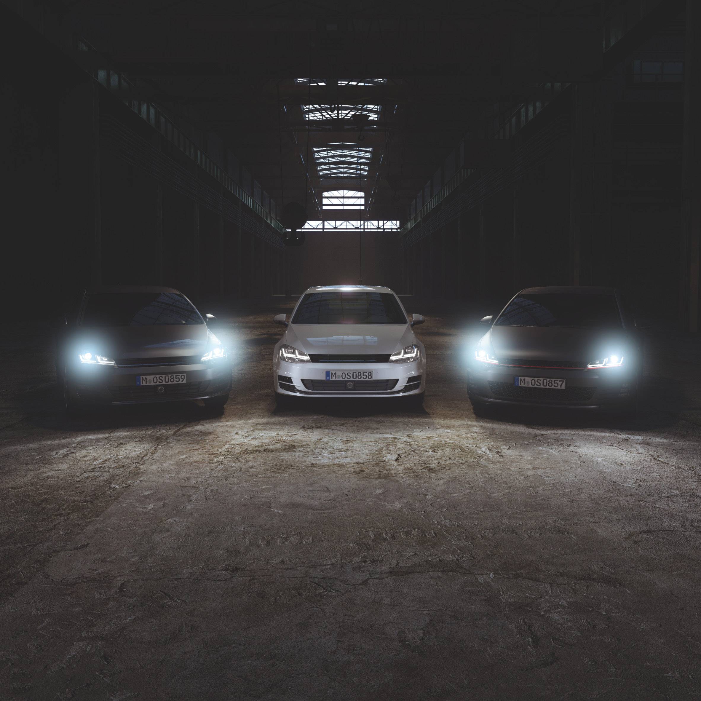 Three cars with headlights on stand side by side in a dark, industrial hall.