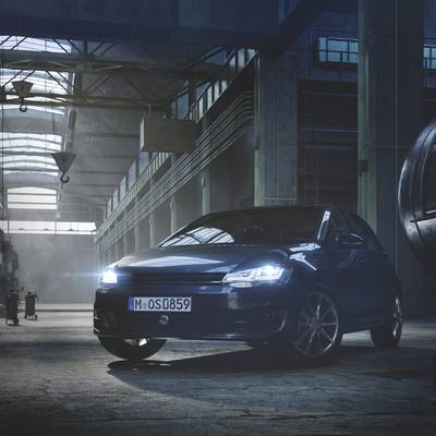 A blue car is parked in a well-lit, industrial hall with high ceilings and large windows. Tools can be seen on the left side.