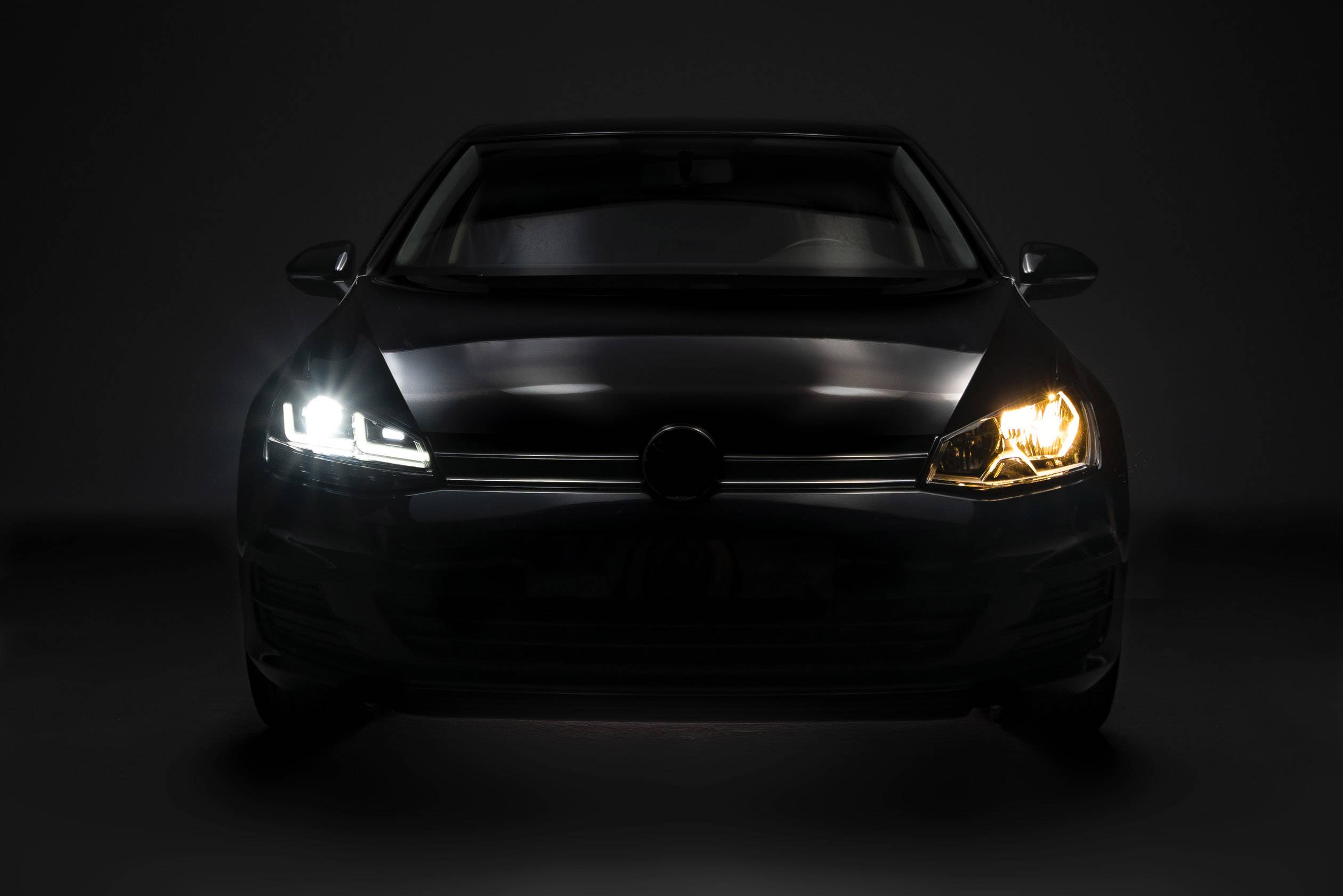 Dark car from the front, headlights switched on, one bright and the other slightly yellowish.