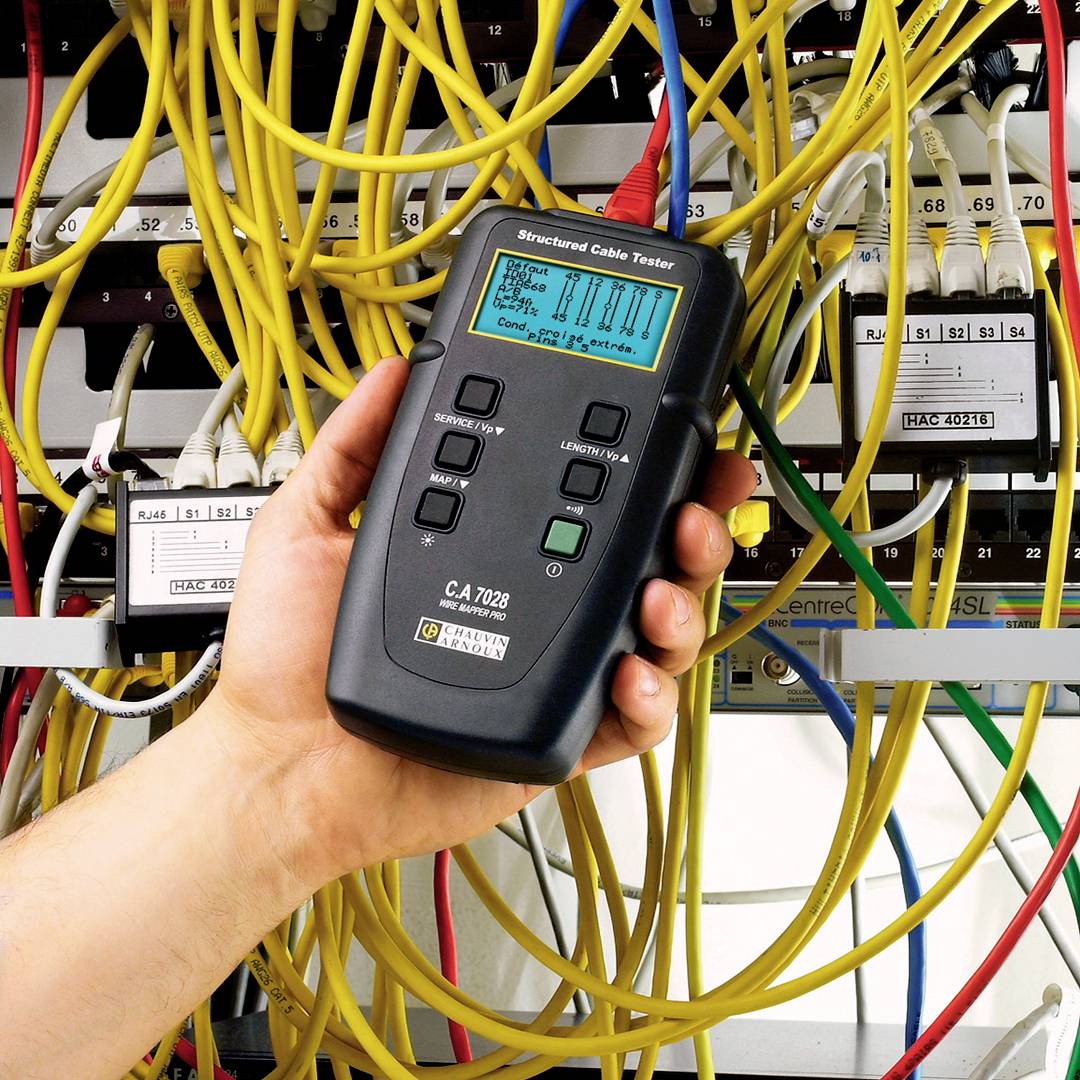 A hand holds a structured cable tester in front of a network distributor with numerous yellow and white cables.