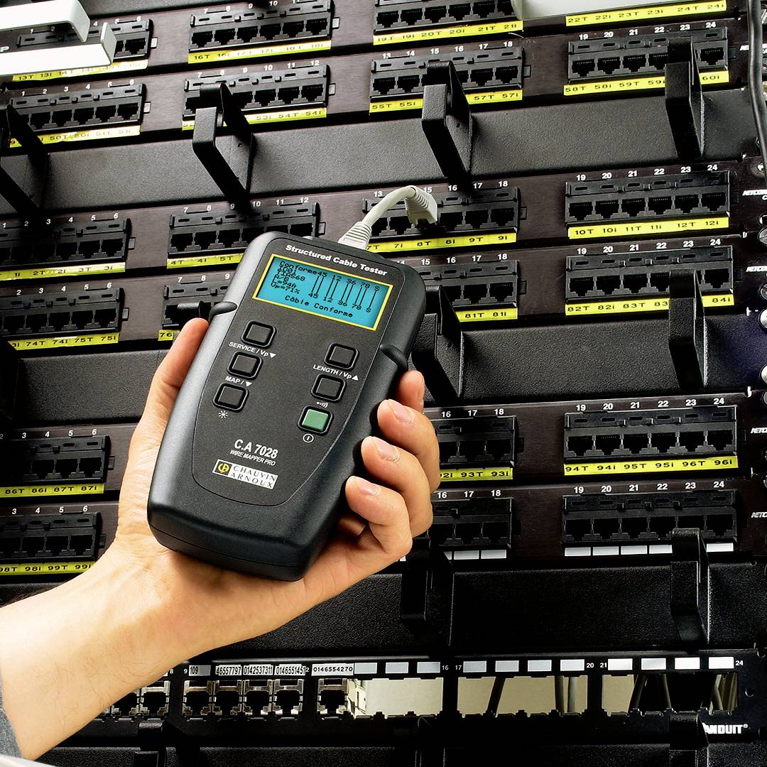 A hand is holding a cable testing device in front of a patch panel. The device displays test results for cable connections on its screen.