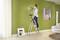 A woman is standing on a ladder and holding a tool while working on a green wall. Artistic picture and lamp as decoration.