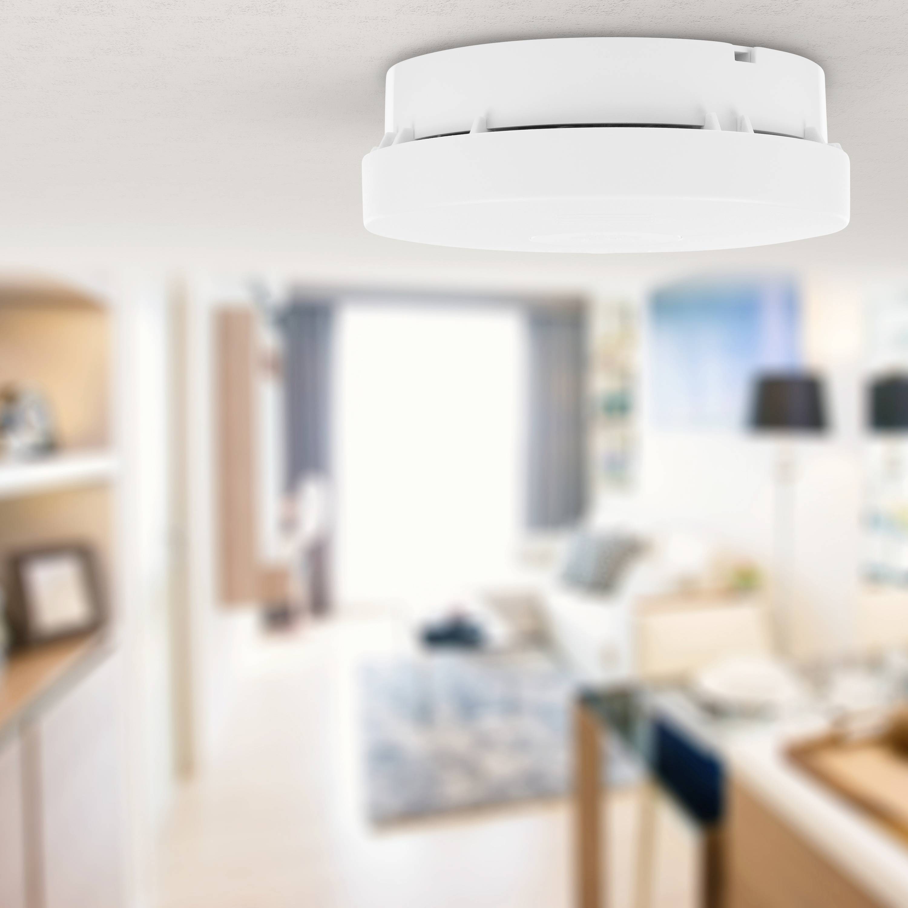 A smoke detector is mounted on the ceiling in a living room. The room is brightly and modernly furnished.