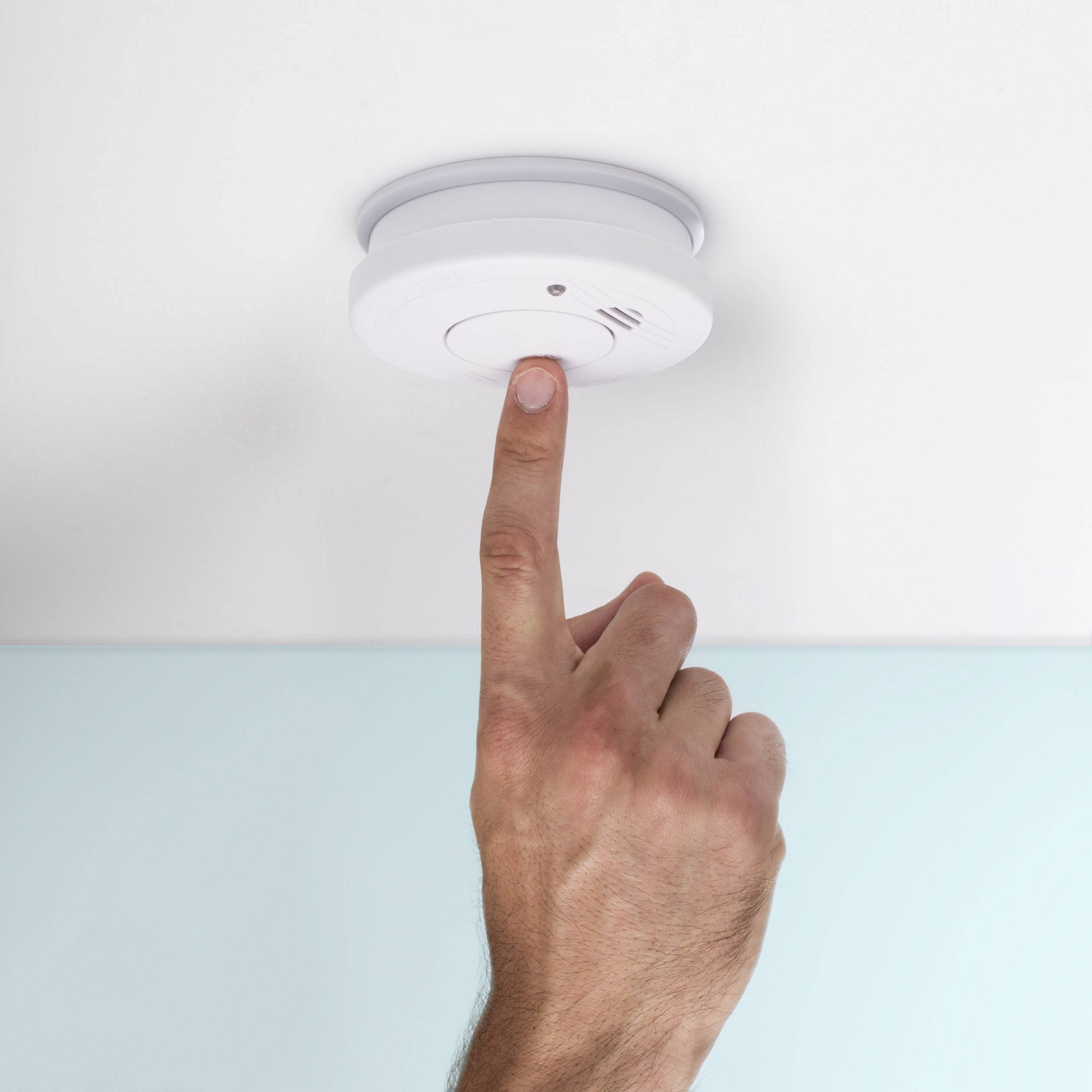 A hand presses the test button on a smoke detector on the ceiling.