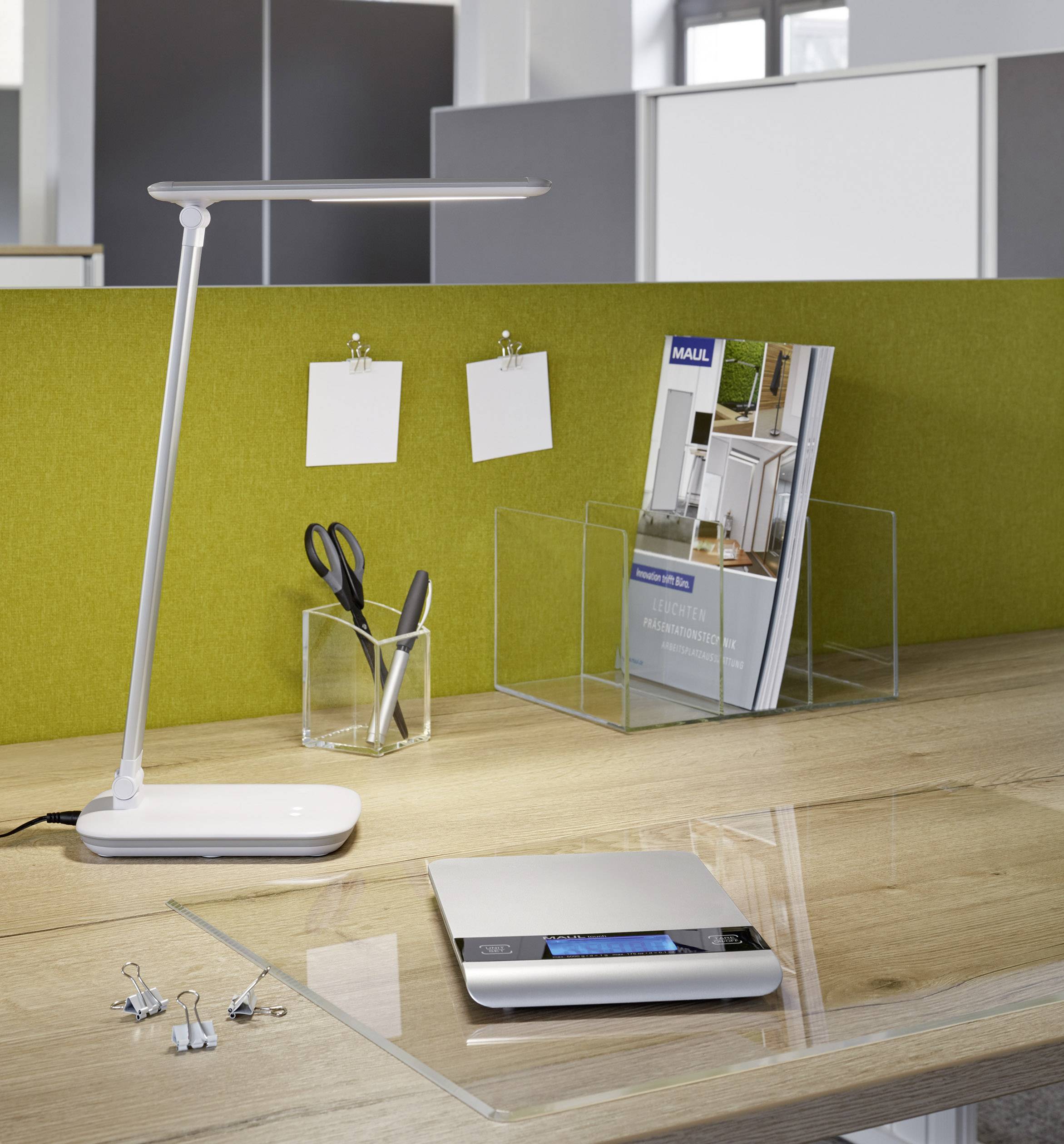 Desk with table lamp, scissors, brochure holder and scales. Background is a green notice board with white sticky notes.