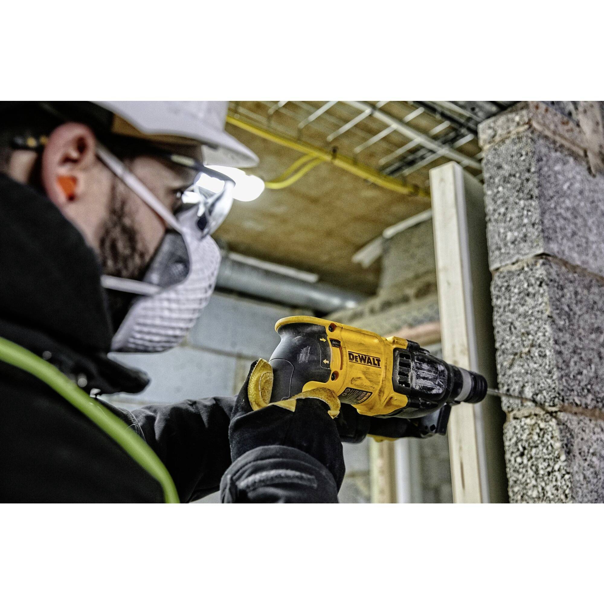 A person wearing protective clothing and a hard hat is drilling a hole in a concrete wall on a construction site using a yellow power drill.