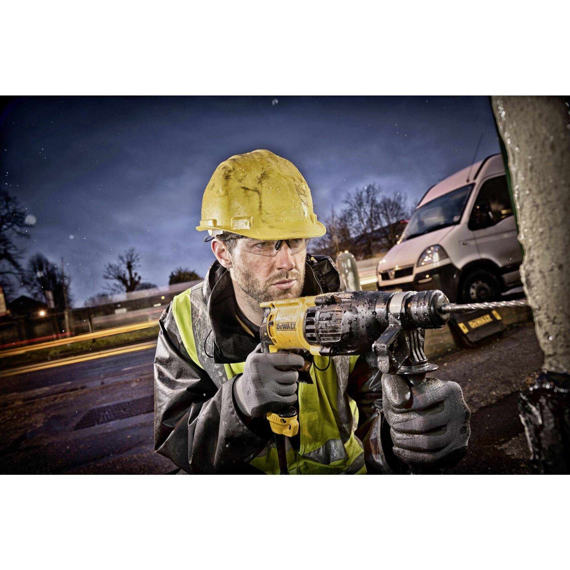 A construction worker wearing a hard hat is drilling with an electric drill outdoors. A van and trees are visible in the background.