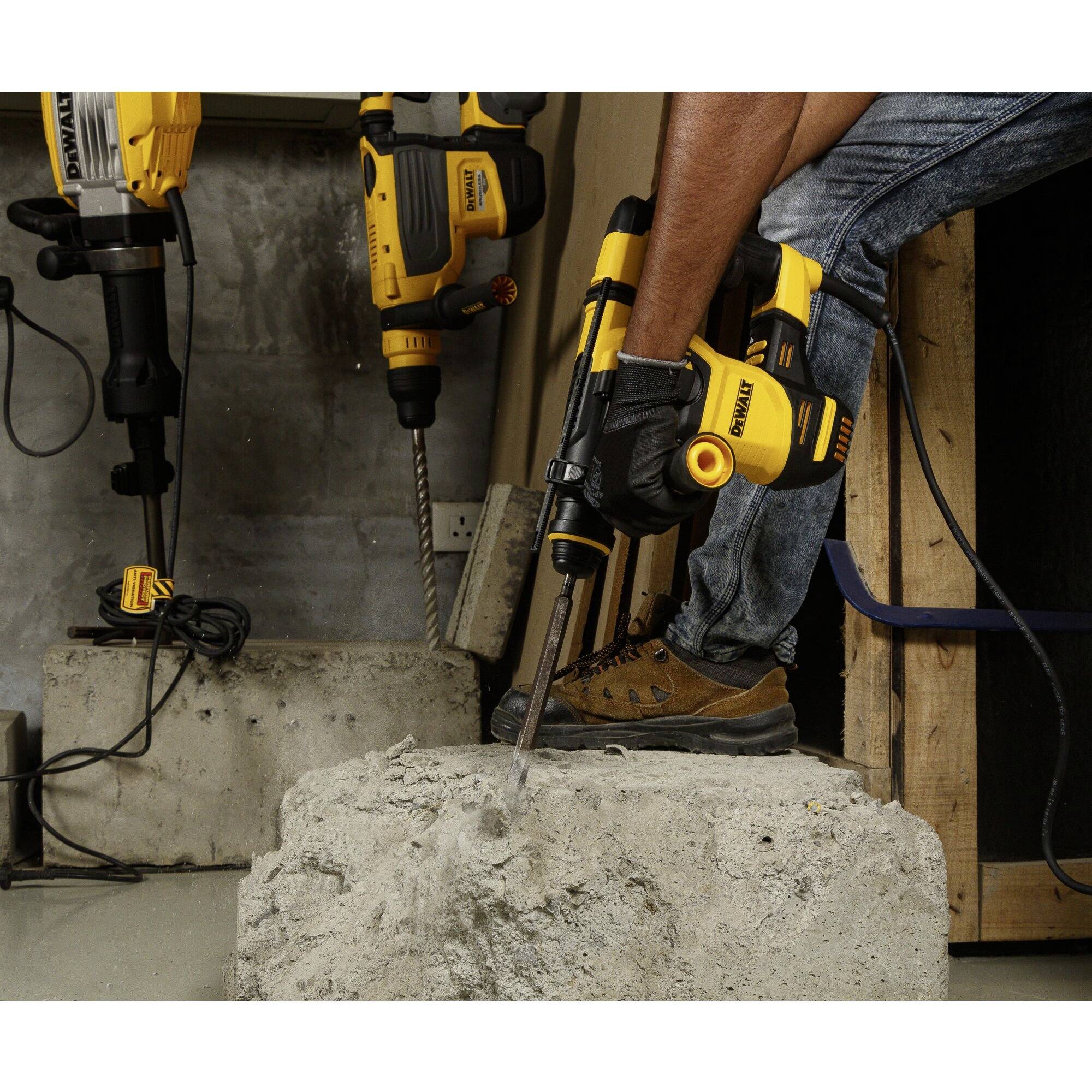 'Man using a drill hammer on a concrete block, two additional drill hammers mounted on the wall; construction site environment in the background.'