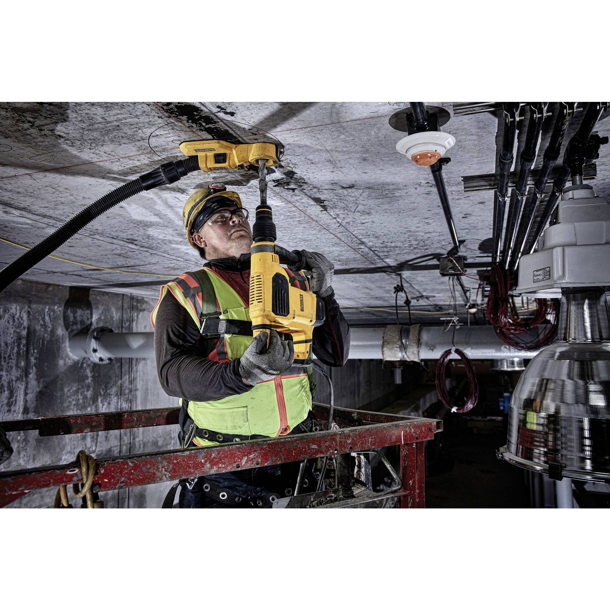 A construction worker on a hydraulic platform is drilling into a concrete ceiling with a yellow drill. He is wearing protective clothing and a hard hat.