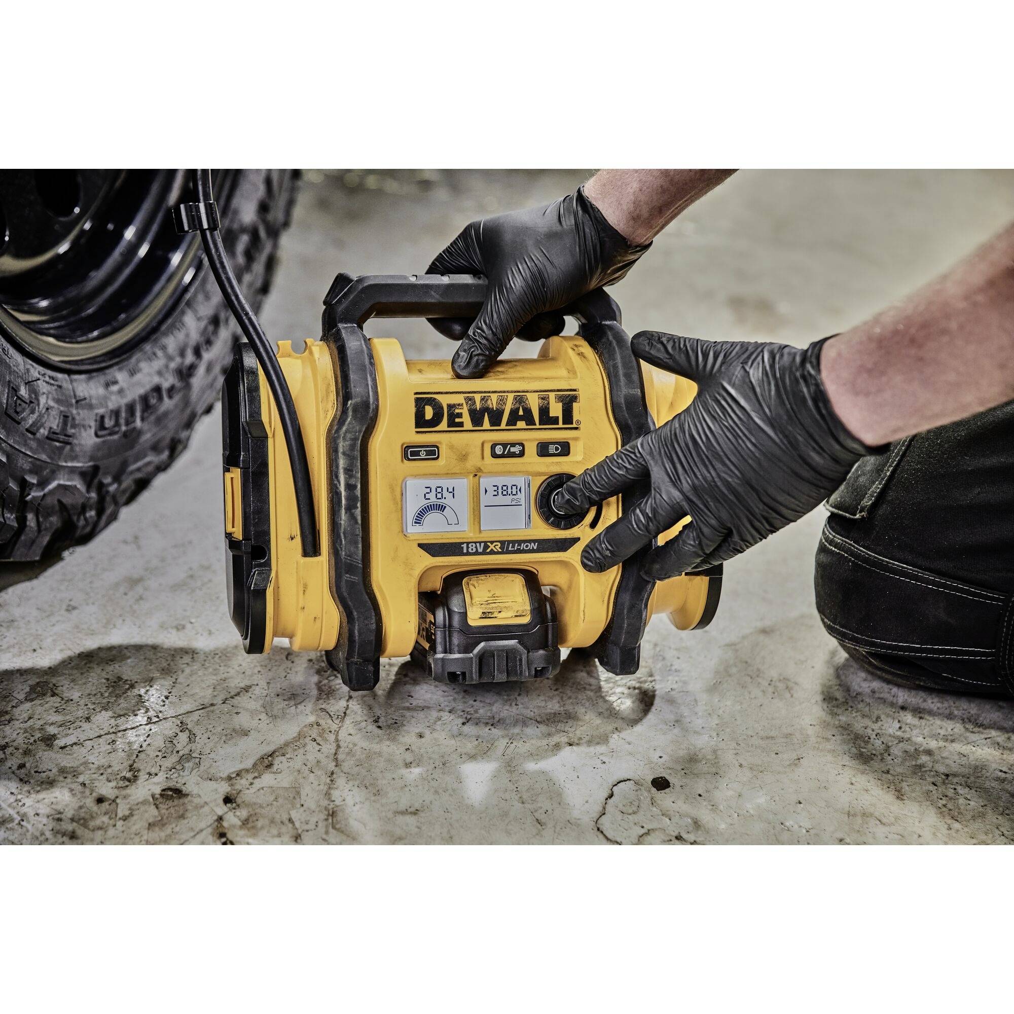 A person wearing black gloves is holding a yellow portable air pump under a vehicle. The device is branded 'DEWALT'.