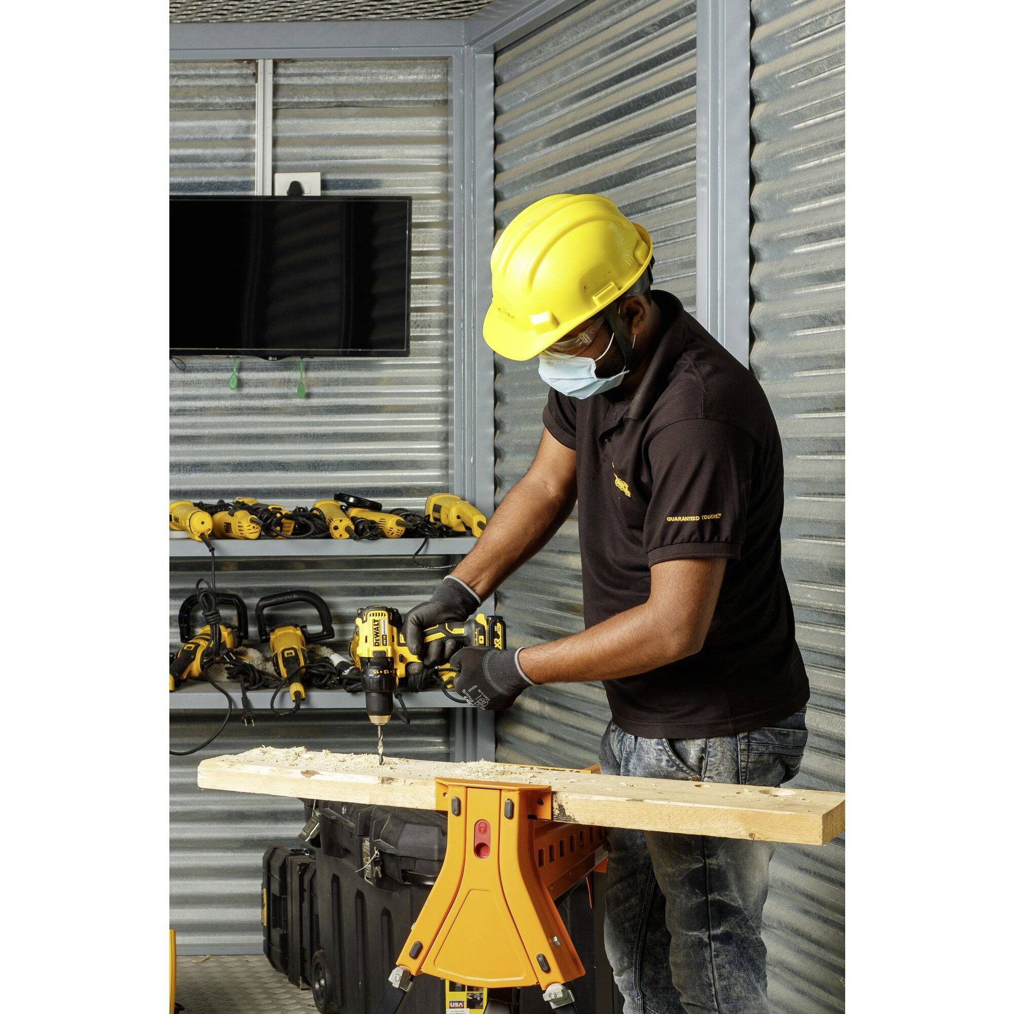 A worker wearing a hard hat and mask is drilling into a wooden board on a workbench. Tools and a monitor are visible in the background.