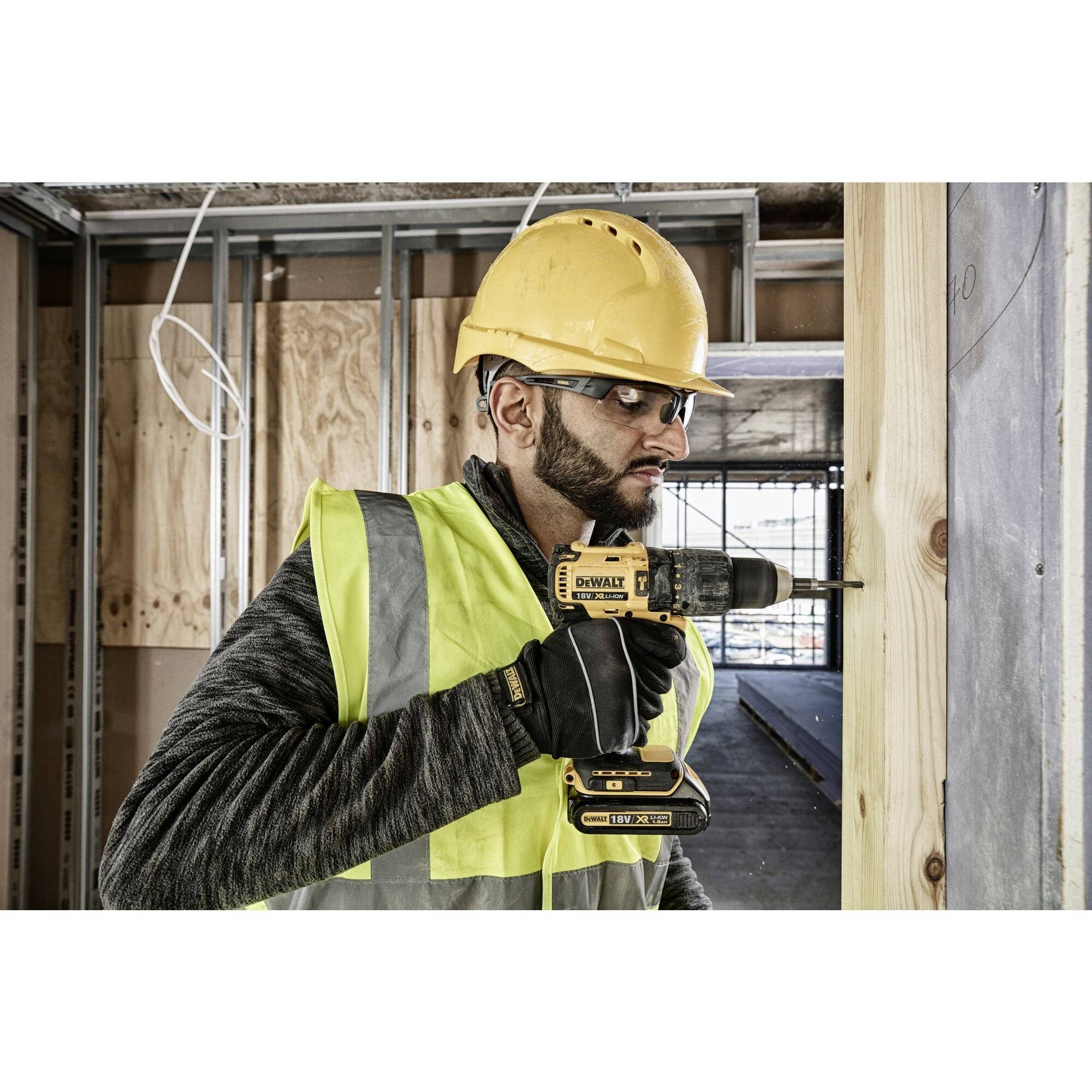A construction worker wearing a hard hat and high-visibility safety vest is using a drill on a wooden post at a building site.