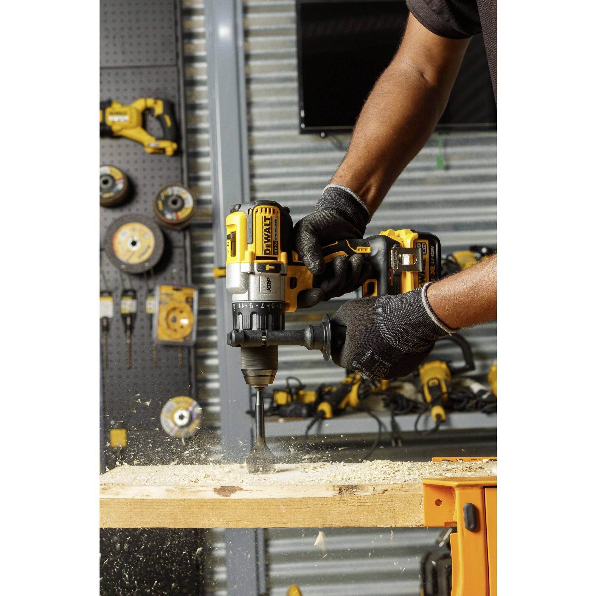 A man is drilling a piece of wood with a yellow drill. Tools are hanging on a wall in the background. Wood shavings are flying around.