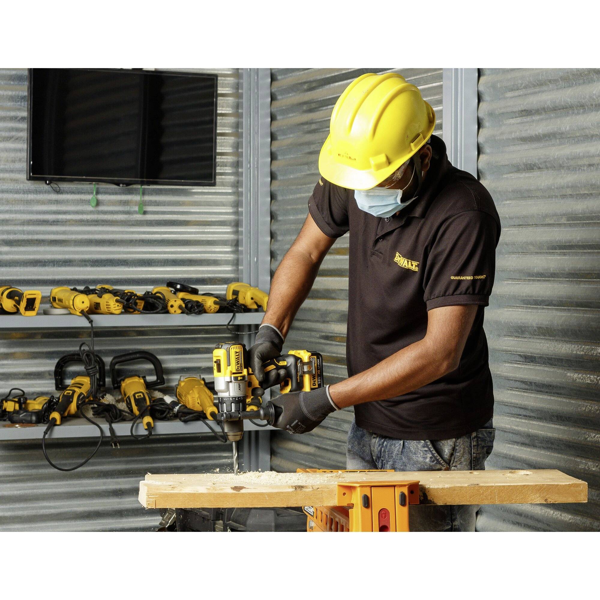 A tradesman wearing a safety helmet and mask is working with an electric tool on a piece of wood, with several tools hanging on the wall in the background.
