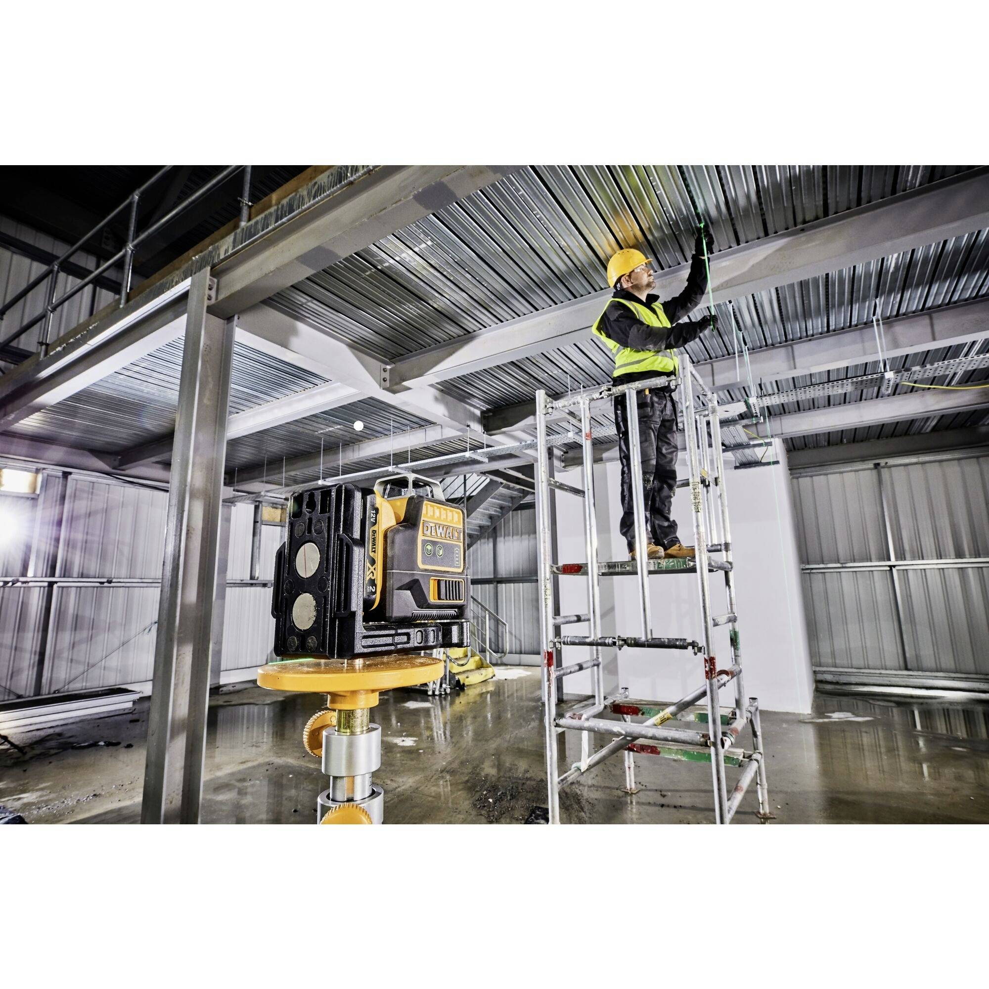 A construction worker is standing on scaffolding in an industrial hall, installing ceiling panels. A laser device is visible in the foreground.