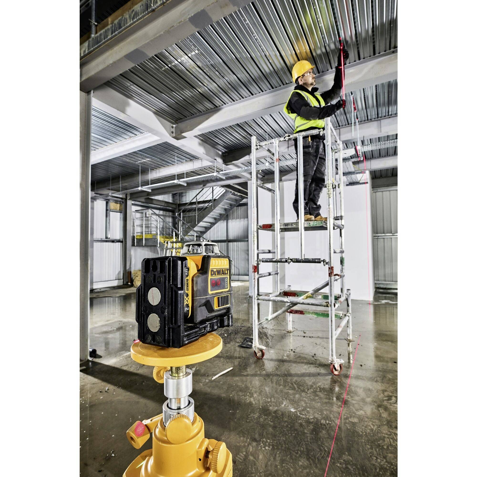 A construction worker stands on scaffolding in a hall and uses a laser device to measure the position of a metal structure on the ceiling.