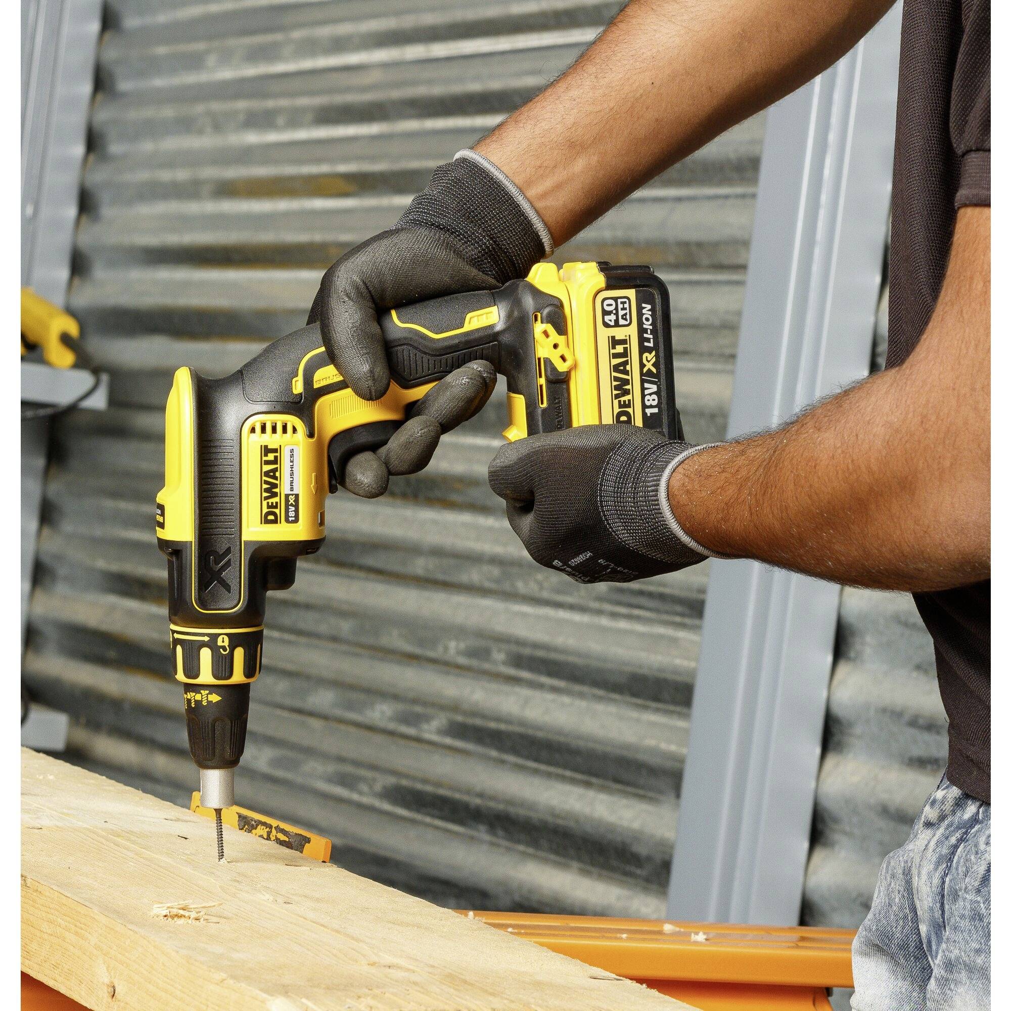 A person is using a yellow cordless drill driver to screw a screw into a wooden board. They are wearing black gloves.