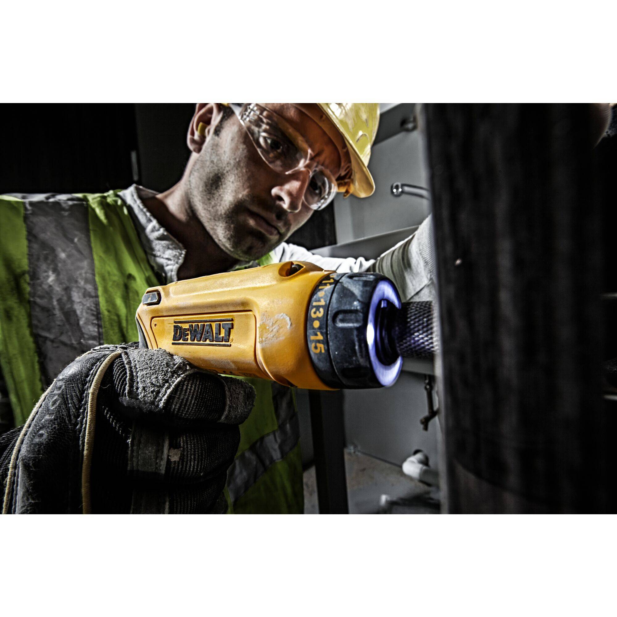 'Worker wearing safety glasses and a hard hat uses a yellow cordless drill, focused on the work in an industrial environment.'