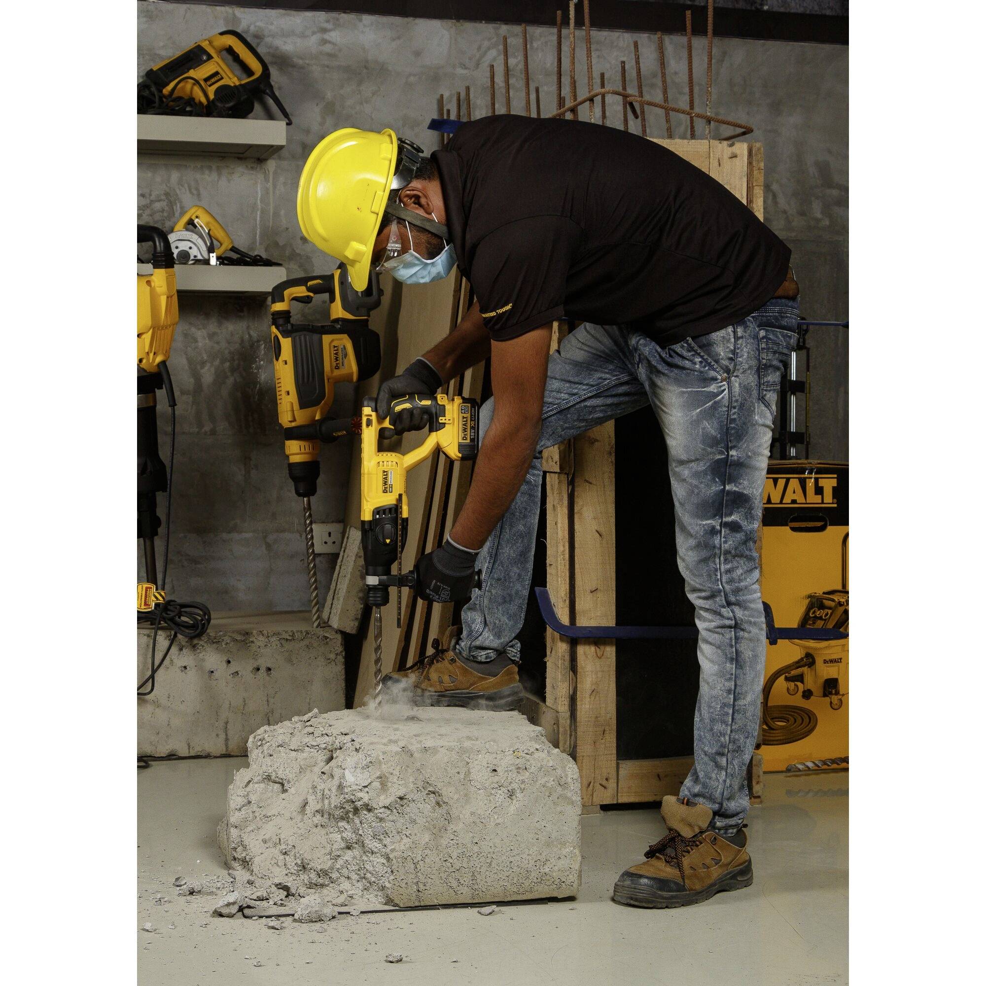 A construction worker wearing a hard hat and mask is drilling into a large concrete block with a yellow drill hammer. Tools can be seen in the background.