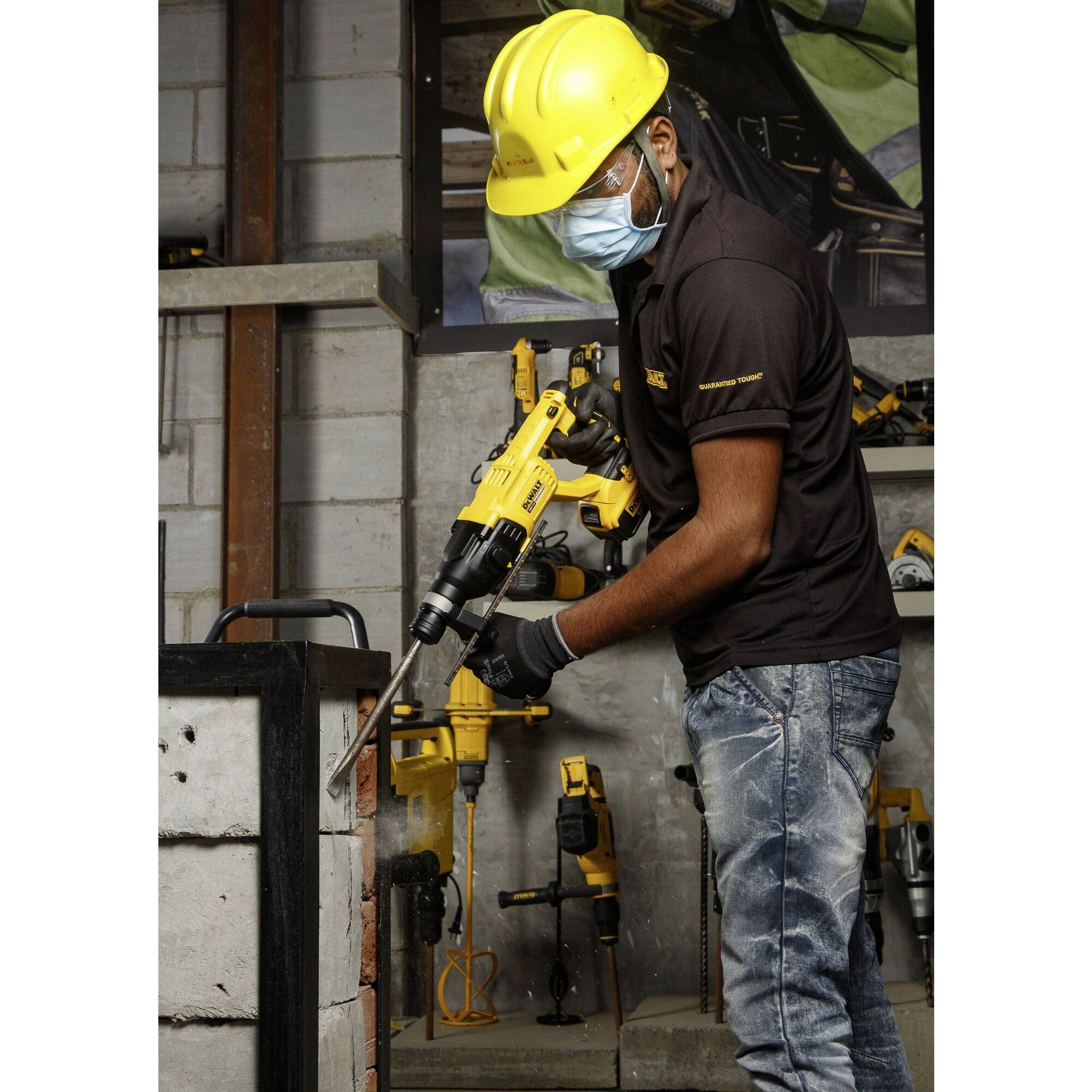 A person wearing a hard hat and mask is working with a drill hammer on a wall in a workshop environment.