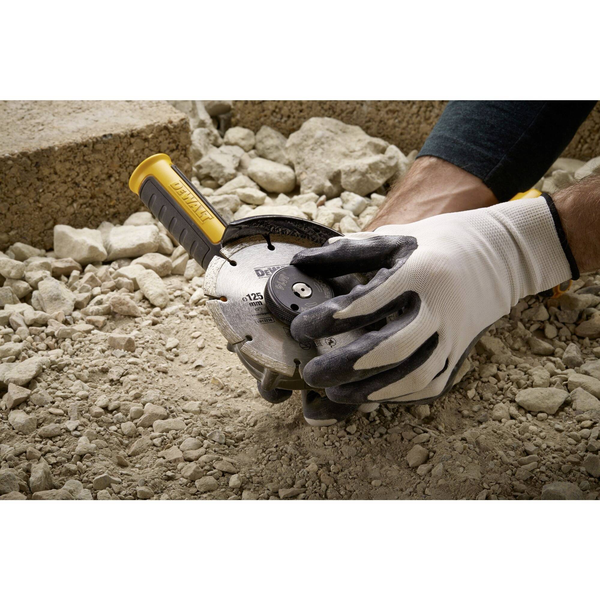 A person is holding an angle grinder, carefully adjusted, on a gravelly surface. They are wearing protective gloves.