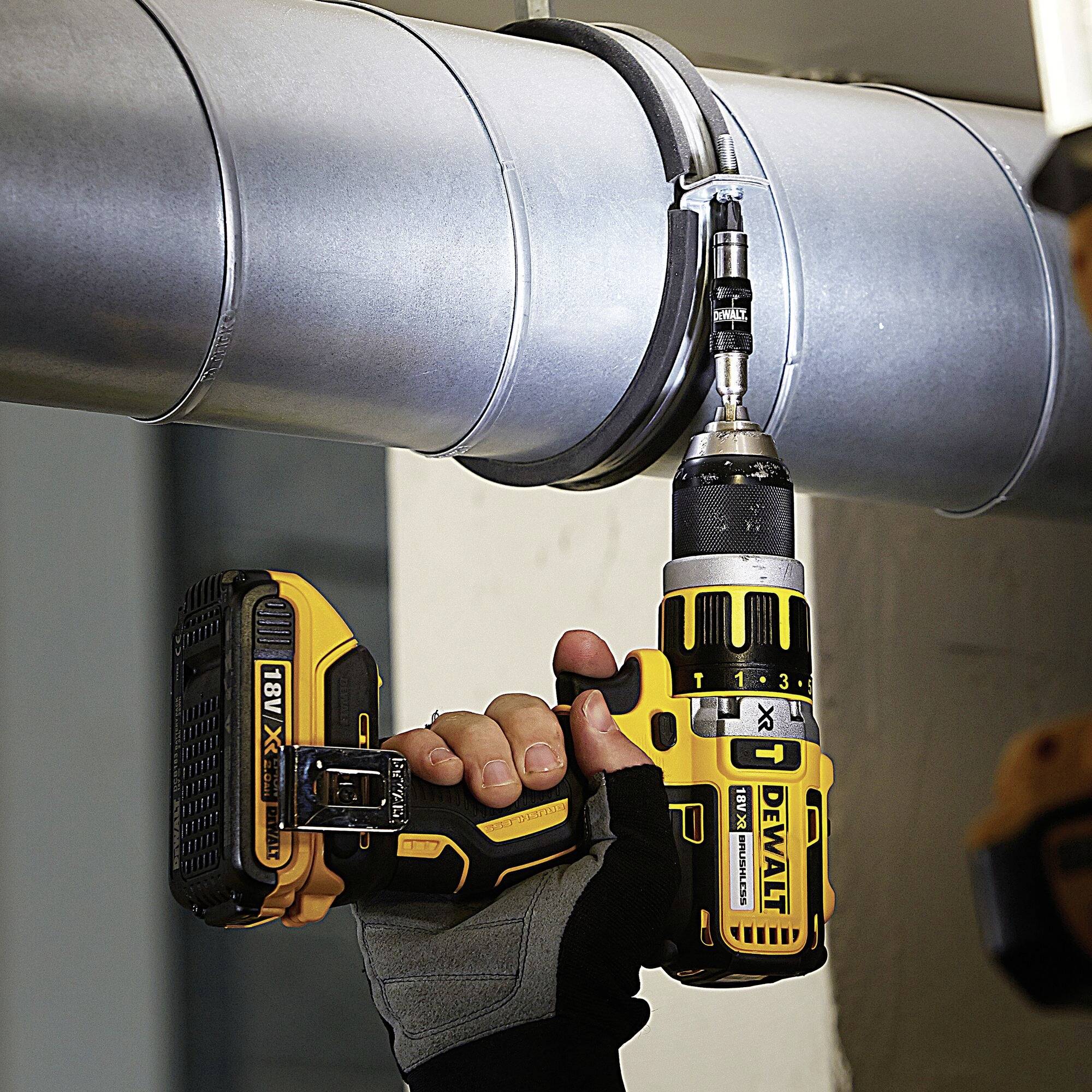 A person is securing a screw to a metal pipe using a cordless drill driver. The tool has a yellow and black handle.