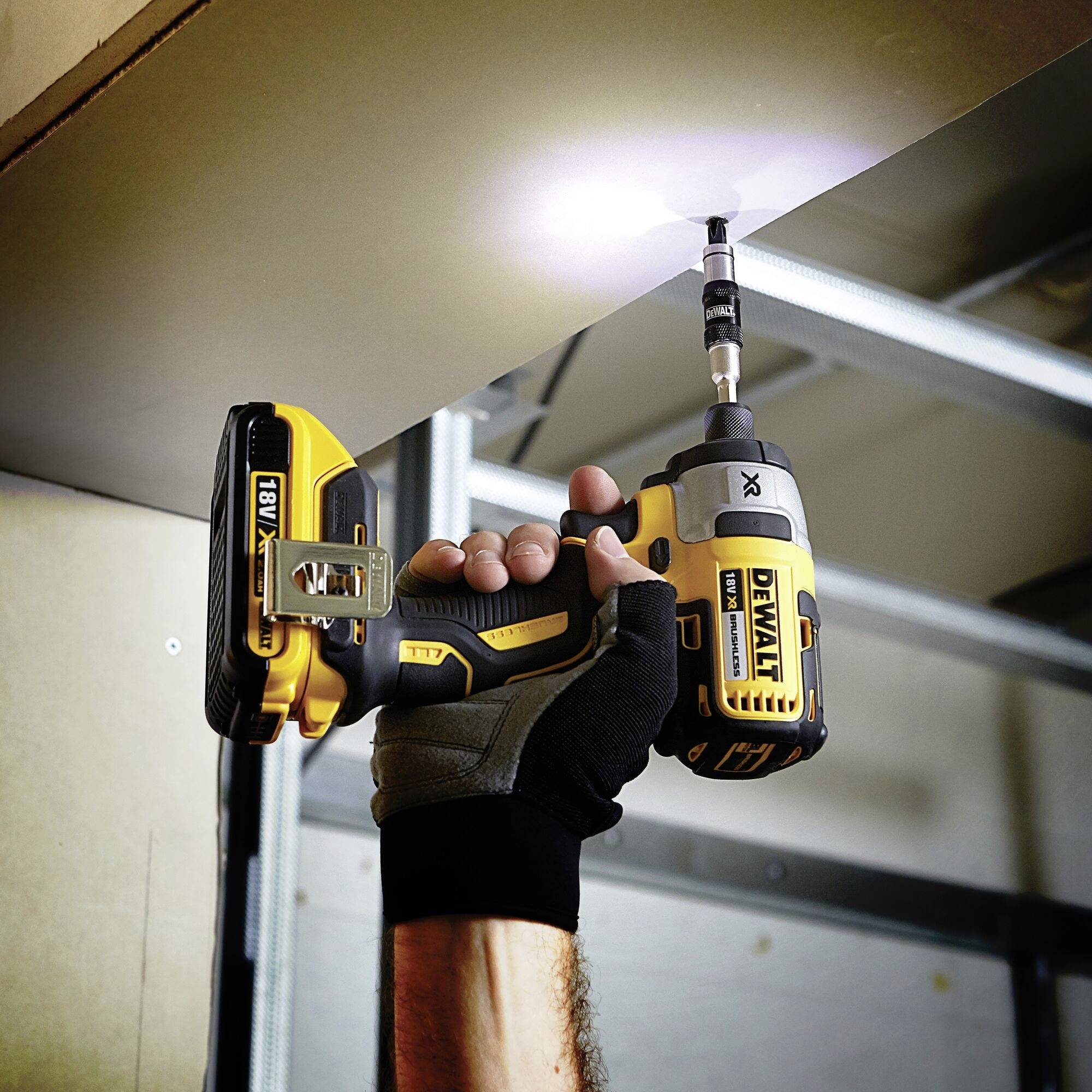 A person is using a yellow cordless drill driver to screw a screw into the ceiling. The environment appears industrial.