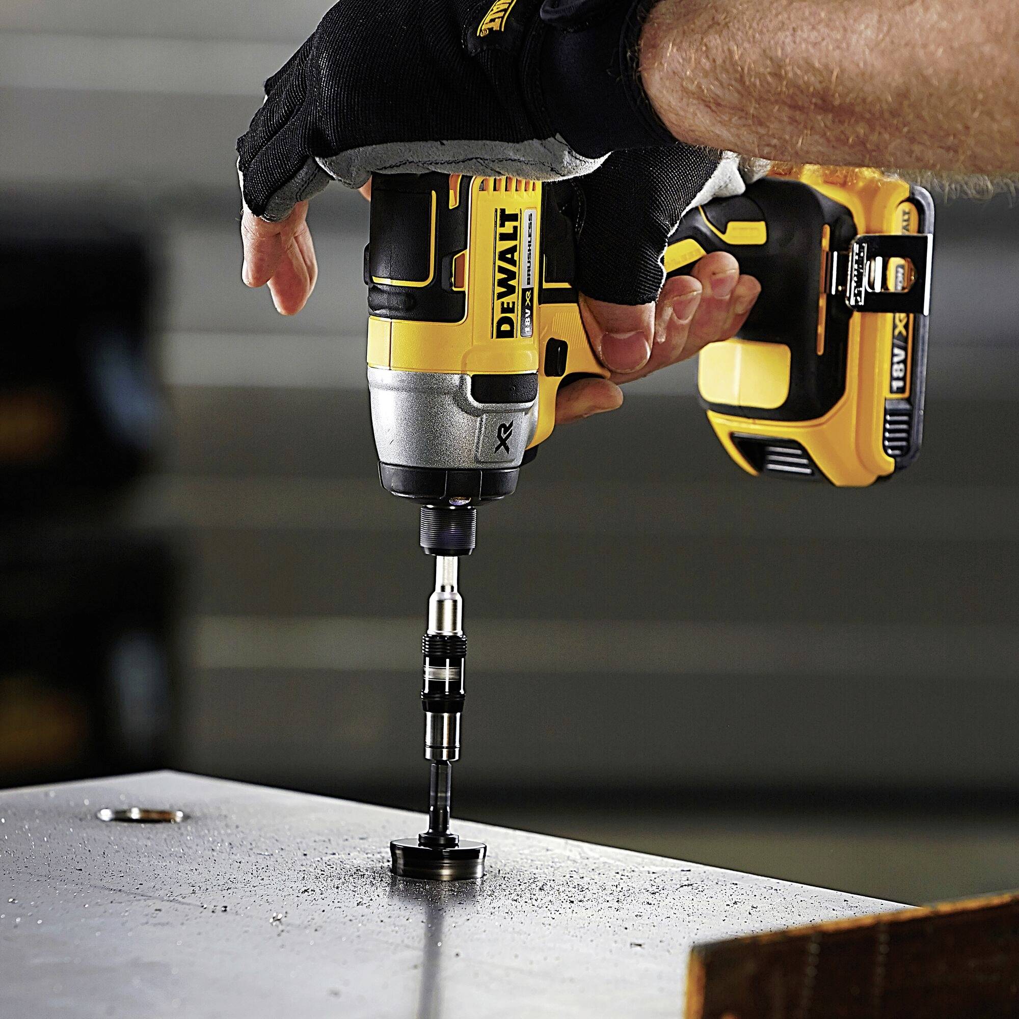 A hand is drilling a hole in a metal plate with a yellow cordless drill, wearing black gloves.