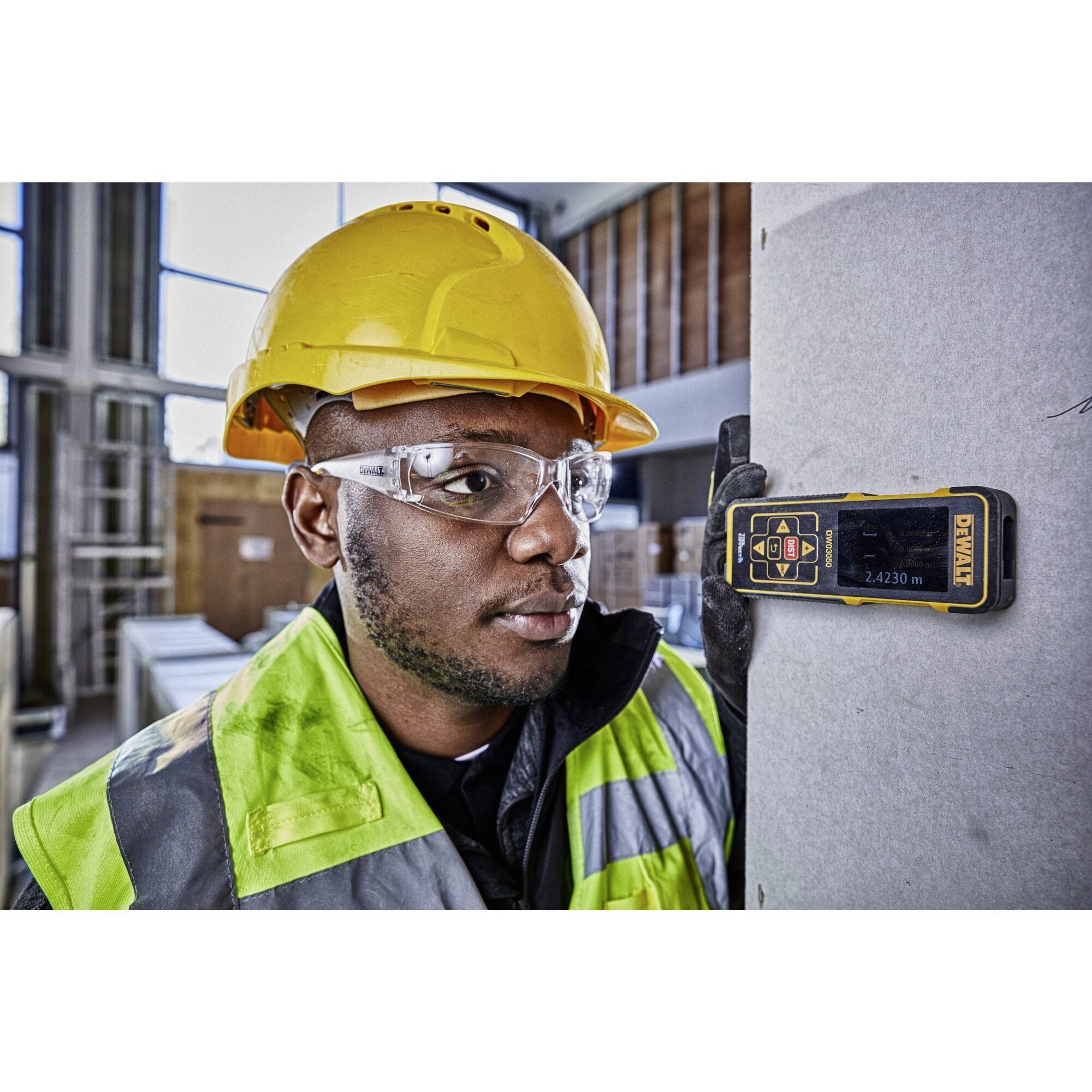 A construction worker wearing a hard hat and safety glasses is measuring a wall with an electronic measuring device in a modern building.