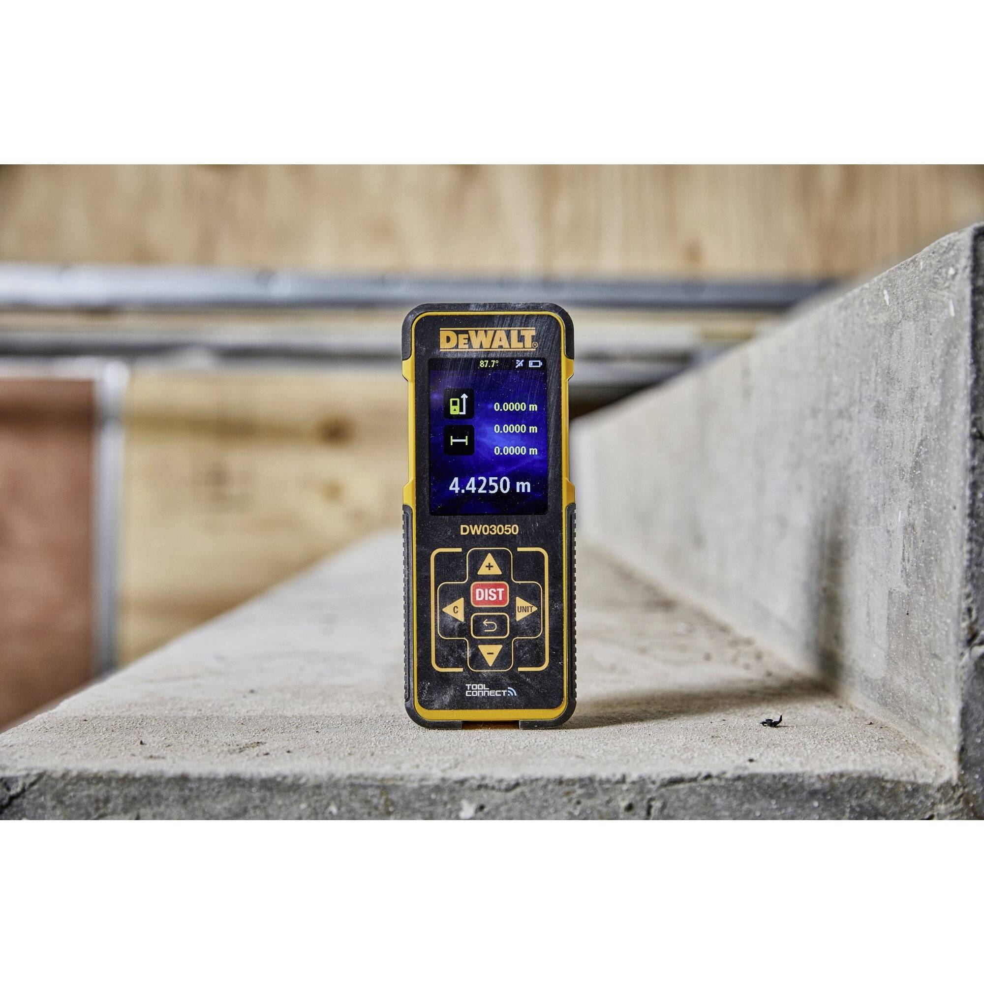 An electronic distance measuring device on a concrete surface displays '4.4250 m' on the screen. Background of wood and metal.