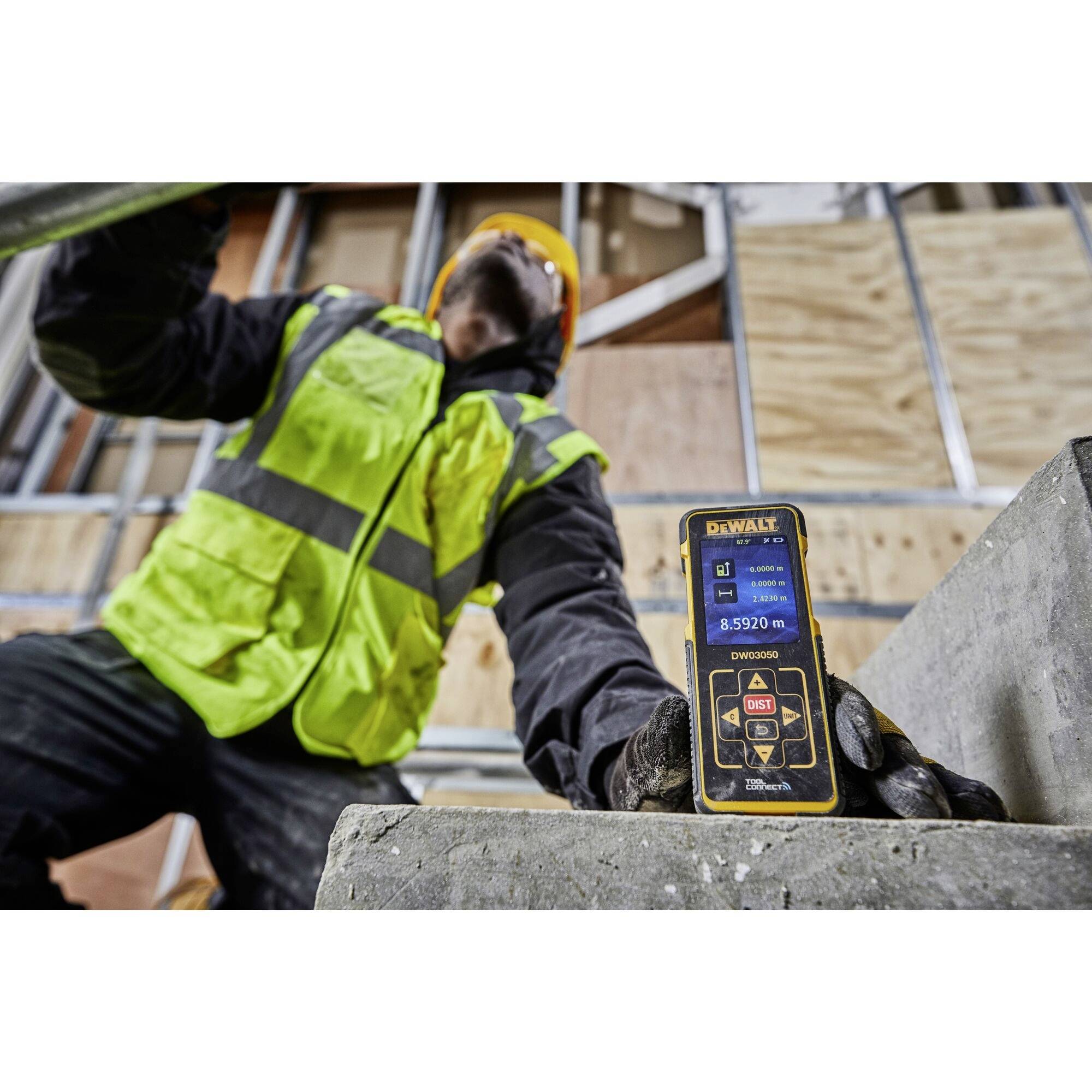 A female construction worker in high-visibility work clothing uses a digital laser distance measuring device in front of an unfinished wall.