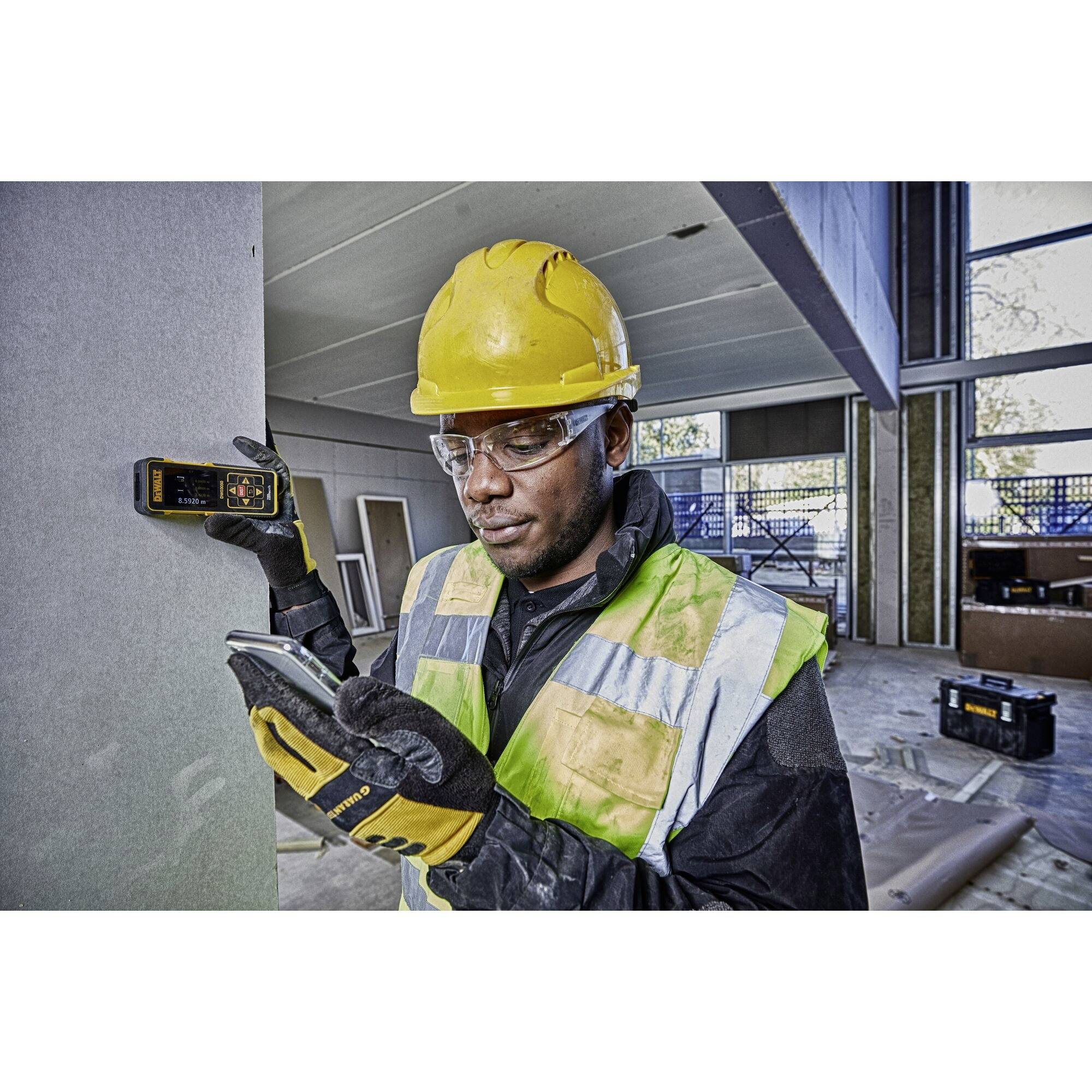 A construction worker wearing a hard hat and high-visibility vest is measuring a wall with a laser tool and noting down results on a smartphone in an unfinished building.