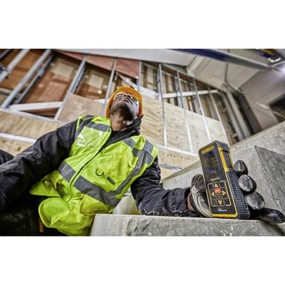 A construction worker wearing a hard hat and high-visibility vest is measuring the distance of a building section using a digital device on a construction site.