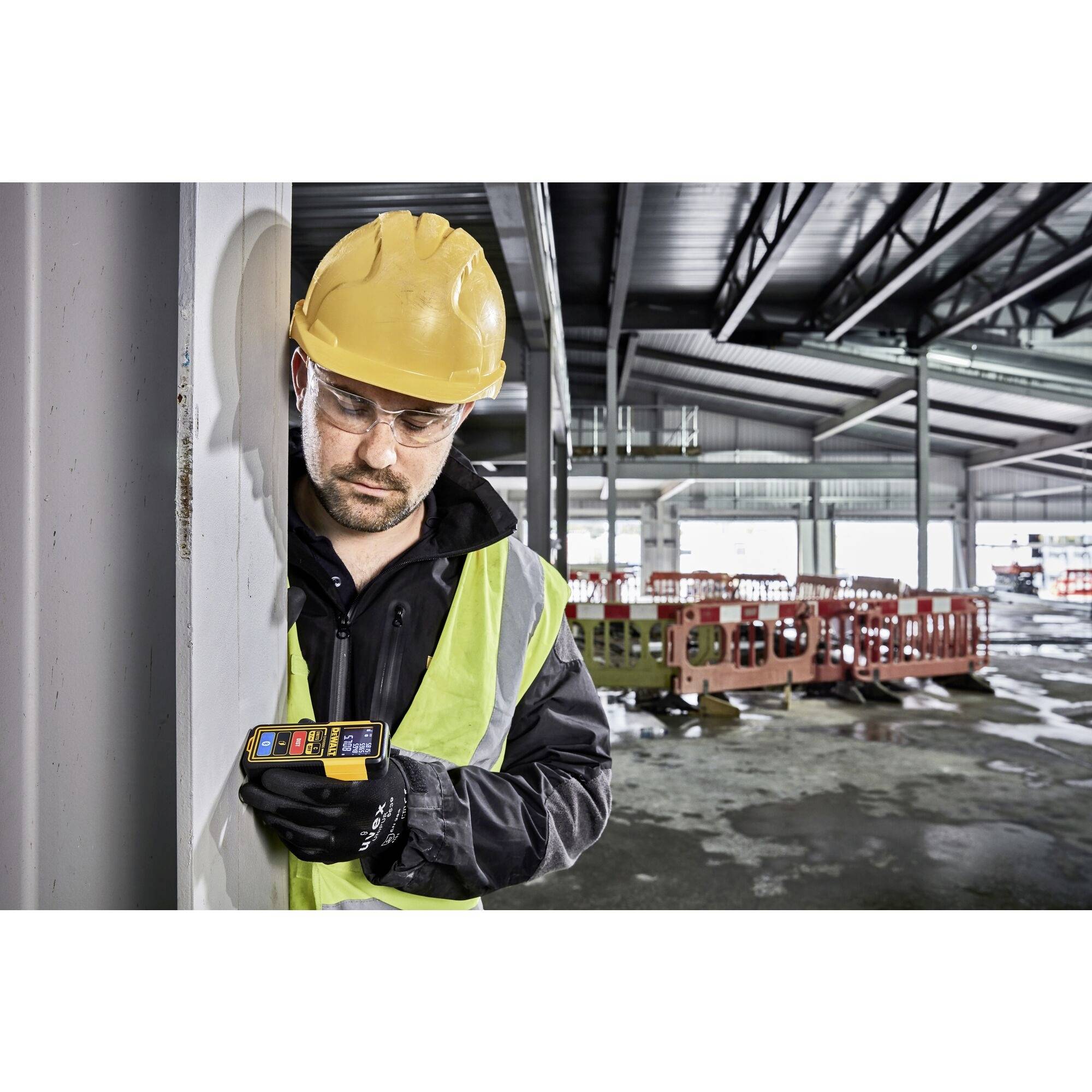 A construction worker in safety clothing and hard hat uses a measuring device on a building site with an incomplete roof and perimeter fencing.