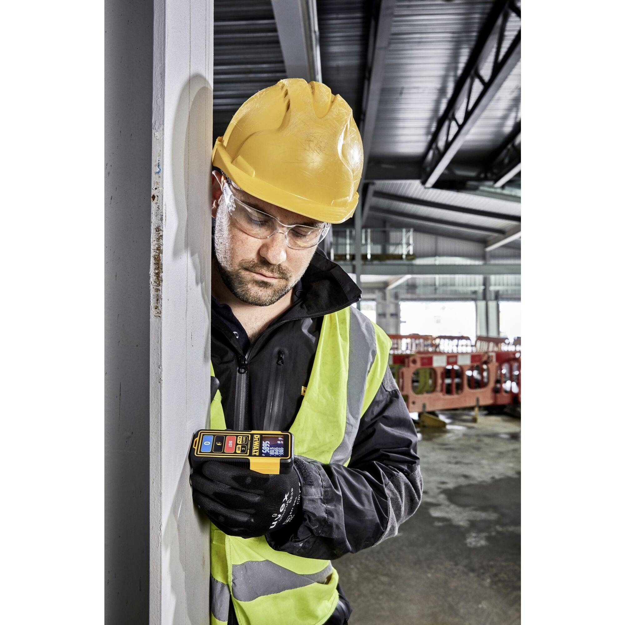 A construction worker wearing a hard hat and high-visibility vest is measuring the distance to a steel post inside an industrial warehouse using a handheld device.