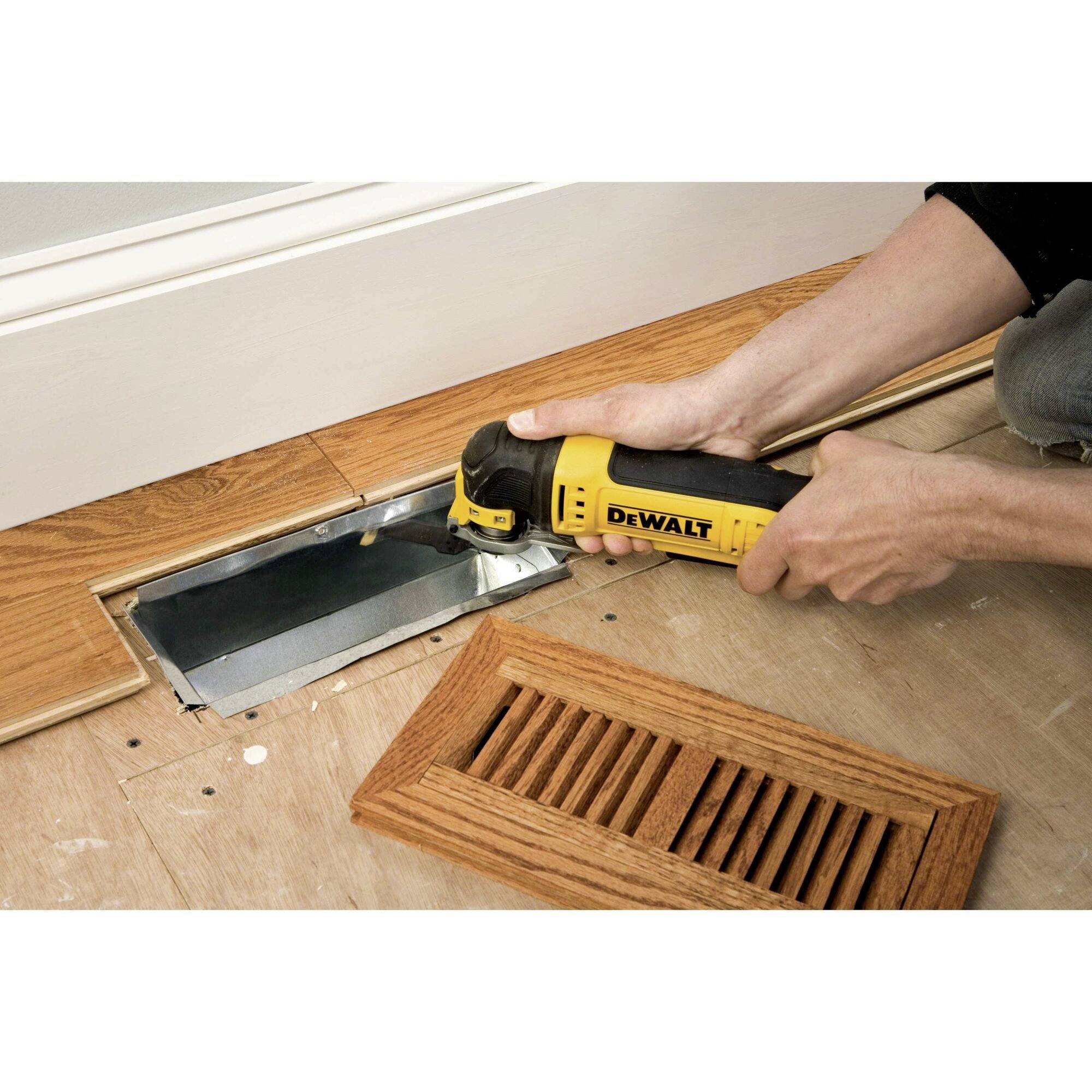 A person is using a yellow power tool to saw a rectangular hole in a wooden floor; a ventilation grille lies nearby.