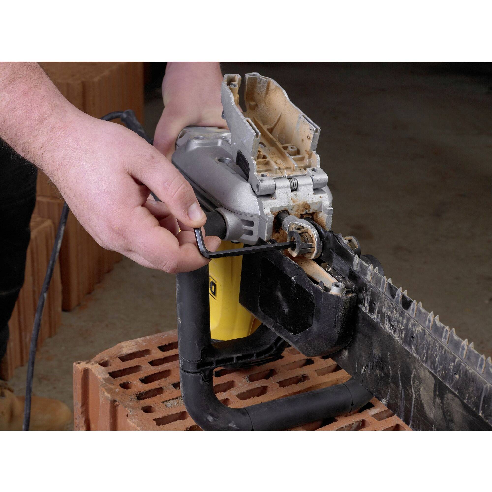 A person is adjusting a chainsaw with a spanner. The saw is resting on a stack of bricks, with a workshop visible in the background.