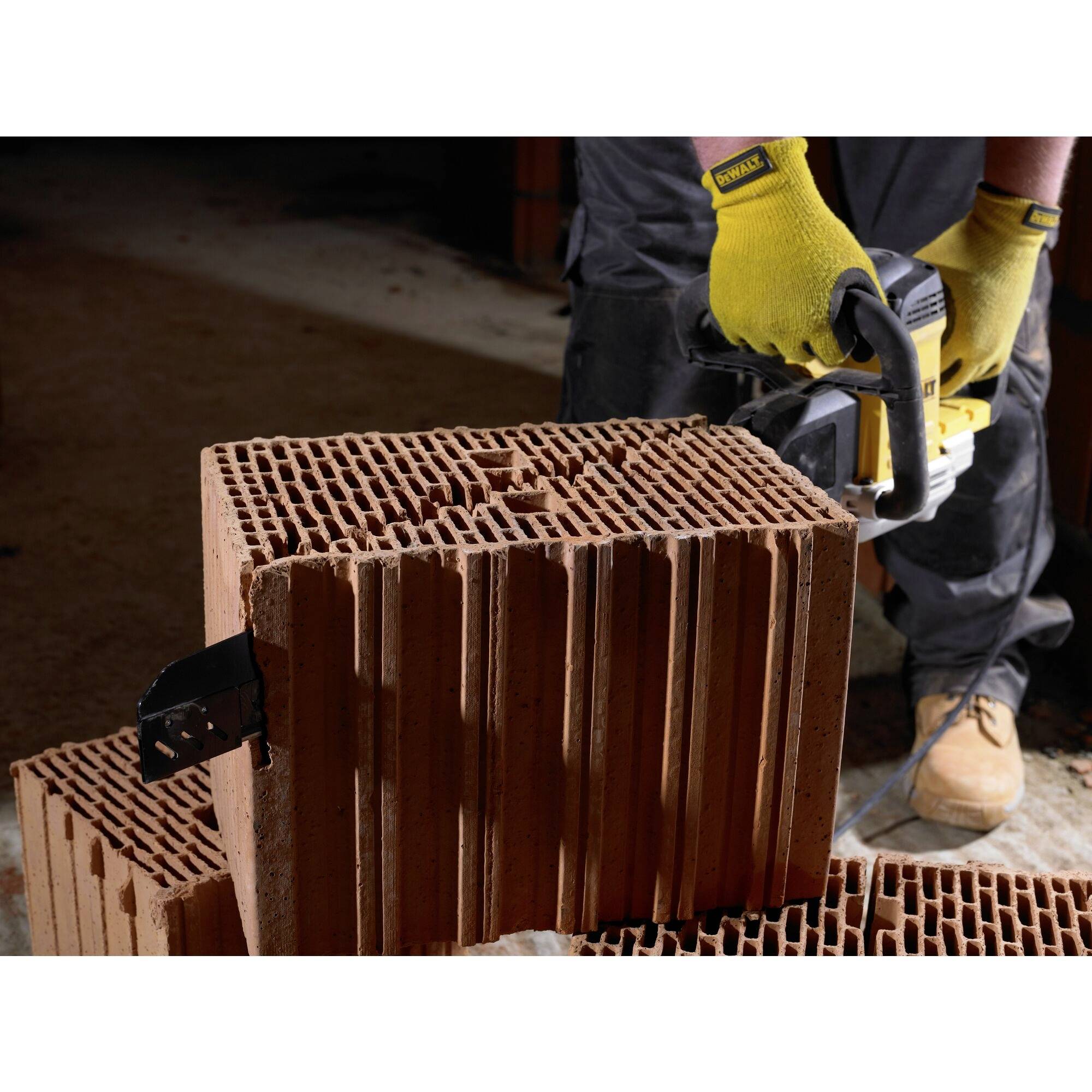 A worker wearing gloves is cutting a large red brick with an electric saw on a construction site.