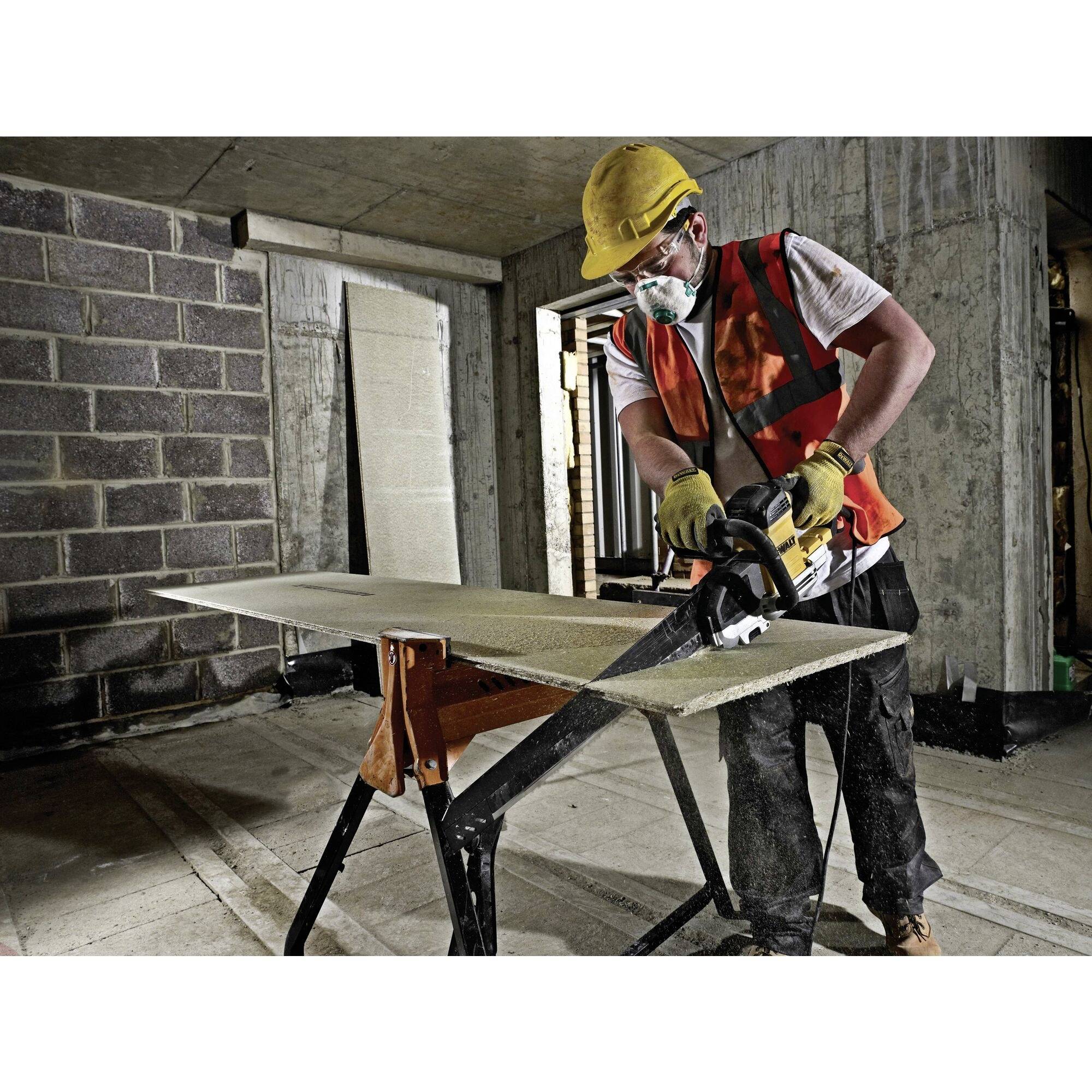 A construction worker wearing a hard hat and mask is sawing a wooden board in an unfinished building. Dust protection clothing is being worn.
